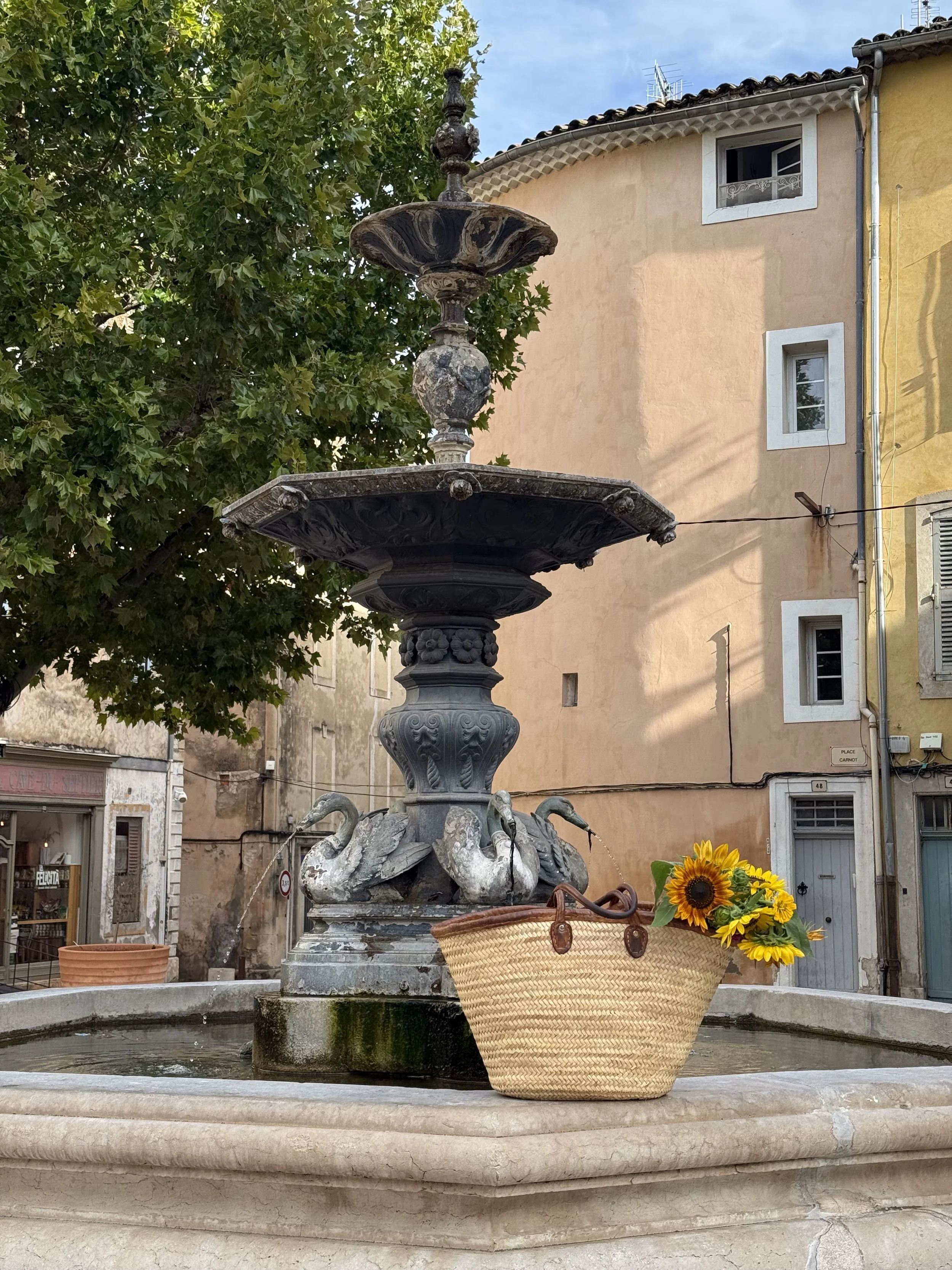 A ornate, multi-tiered stone fountain in a plaza, adorned with swan sculptures and a sunflower arrangement in a woven basket, with trees and buildings in the background.