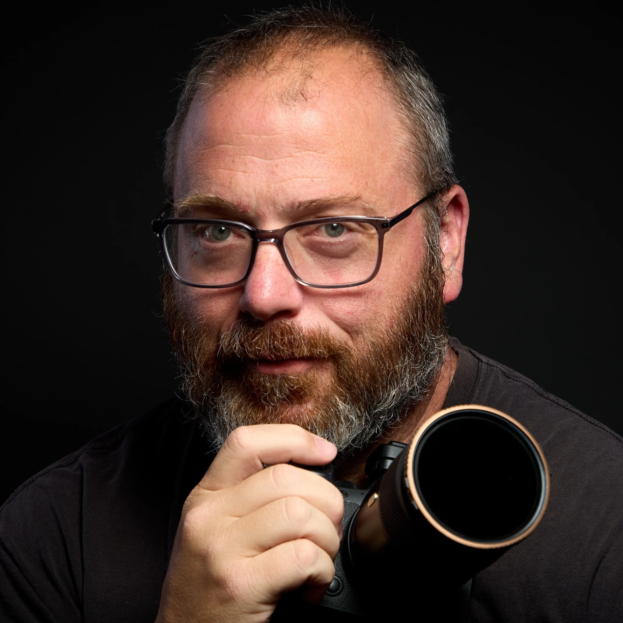 A man with glasses and a beard holding a camera close to his face, partially visible against a black background.