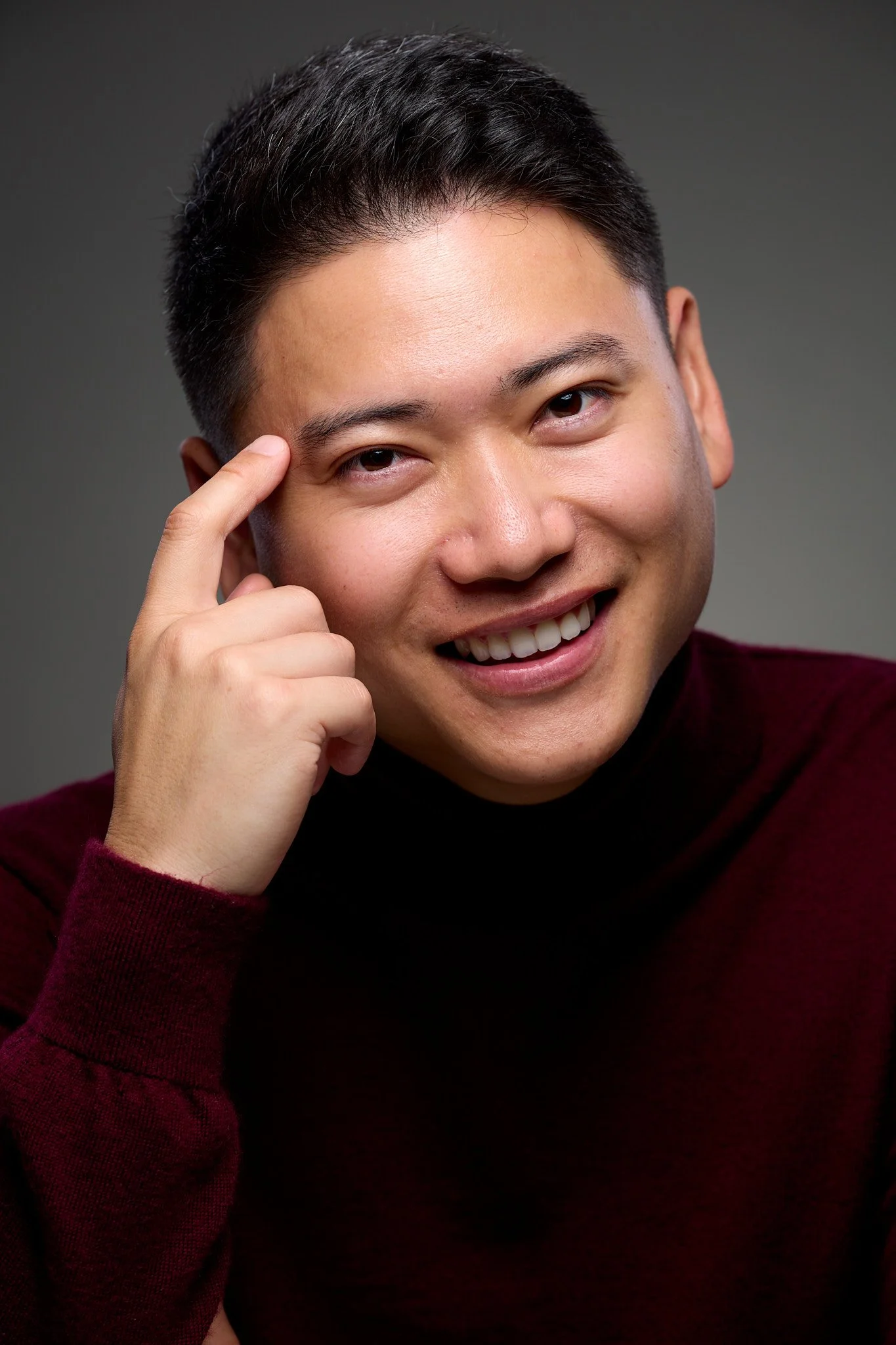 A smiling young man with short dark hair, wearing a maroon sweater, touching his temple with his right hand against a gray gradient background.