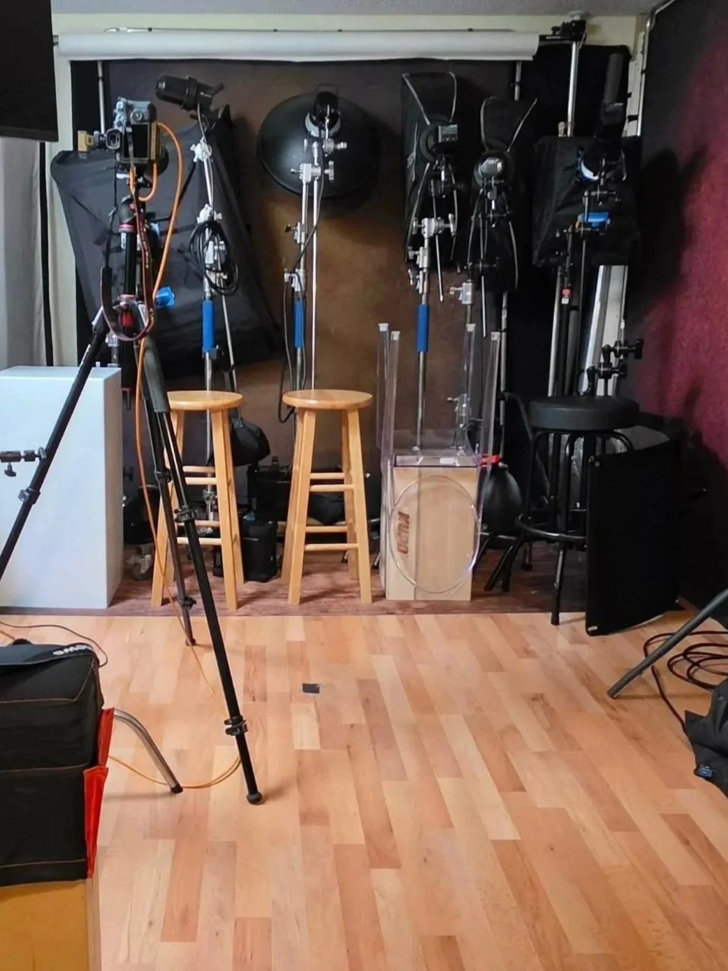 Photography studio setup with various lighting equipment, tripods, stools, and backdrop in a room with wooden flooring.