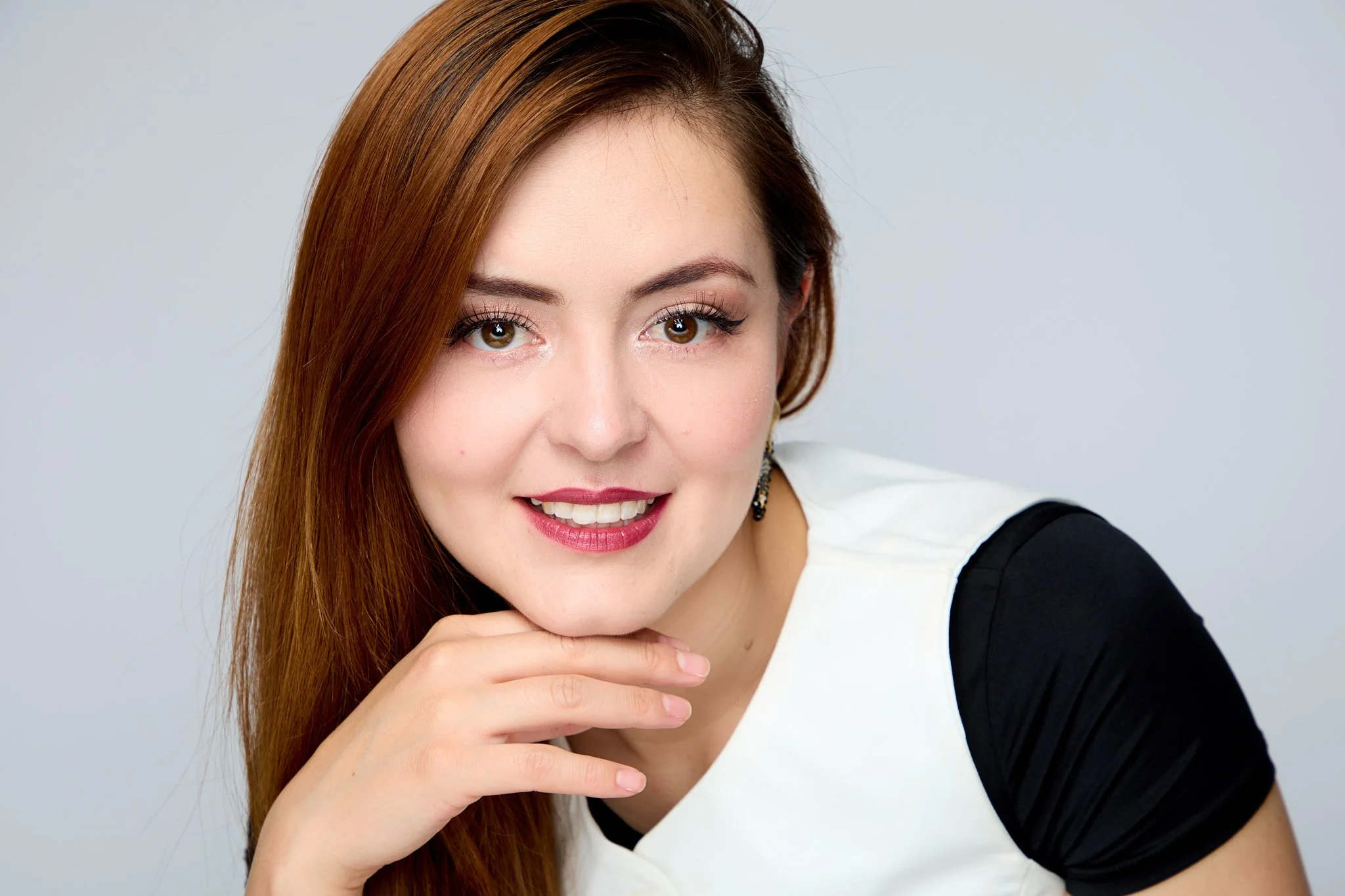 Close-up of a young woman with long reddish-brown hair, smiling, wearing a white shirt with black sleeves, resting her chin on her right hand, against a plain light gray background.