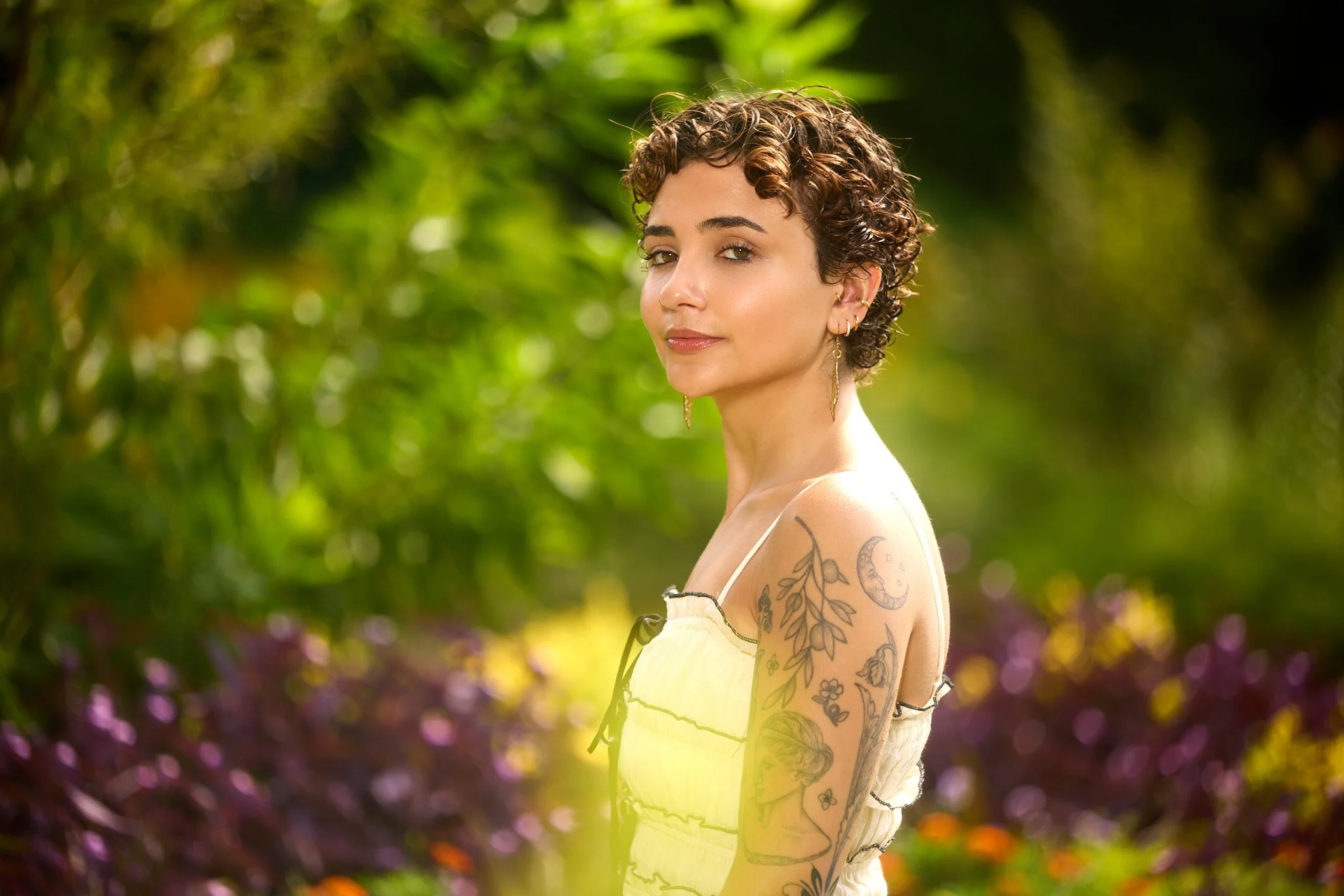 A woman with short curly hair, multiple tattoos on her arm, wearing a yellow dress, standing outdoors with a blurred green and purple floral background.