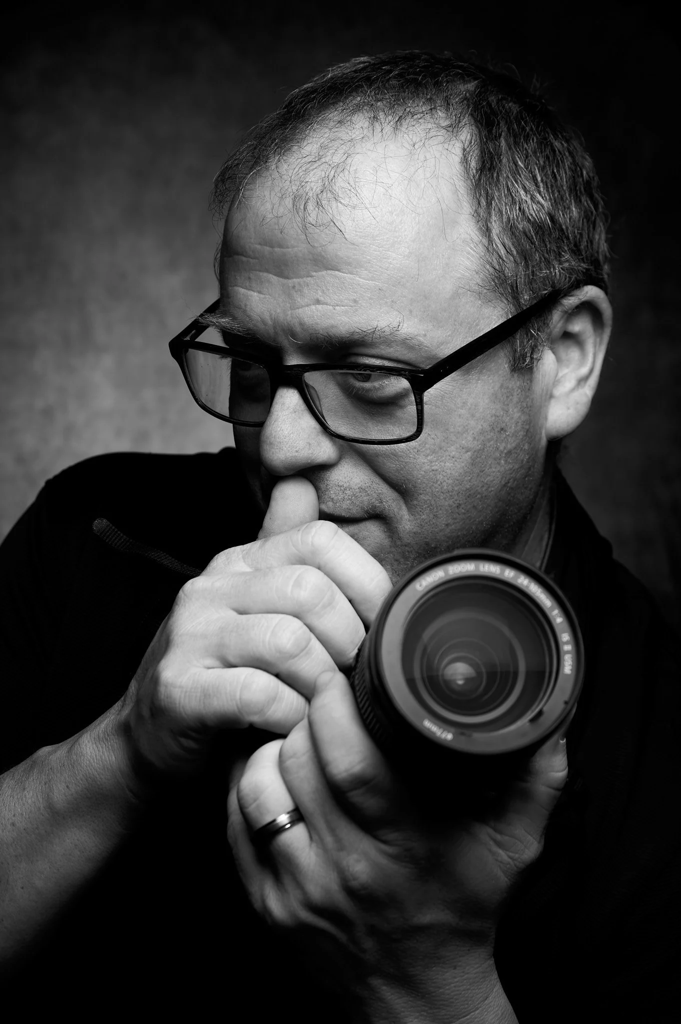 Black and white photo of a man with glasses taking a selfie, holding a camera in front of his face with his right hand, and touching his nose with his left hand.