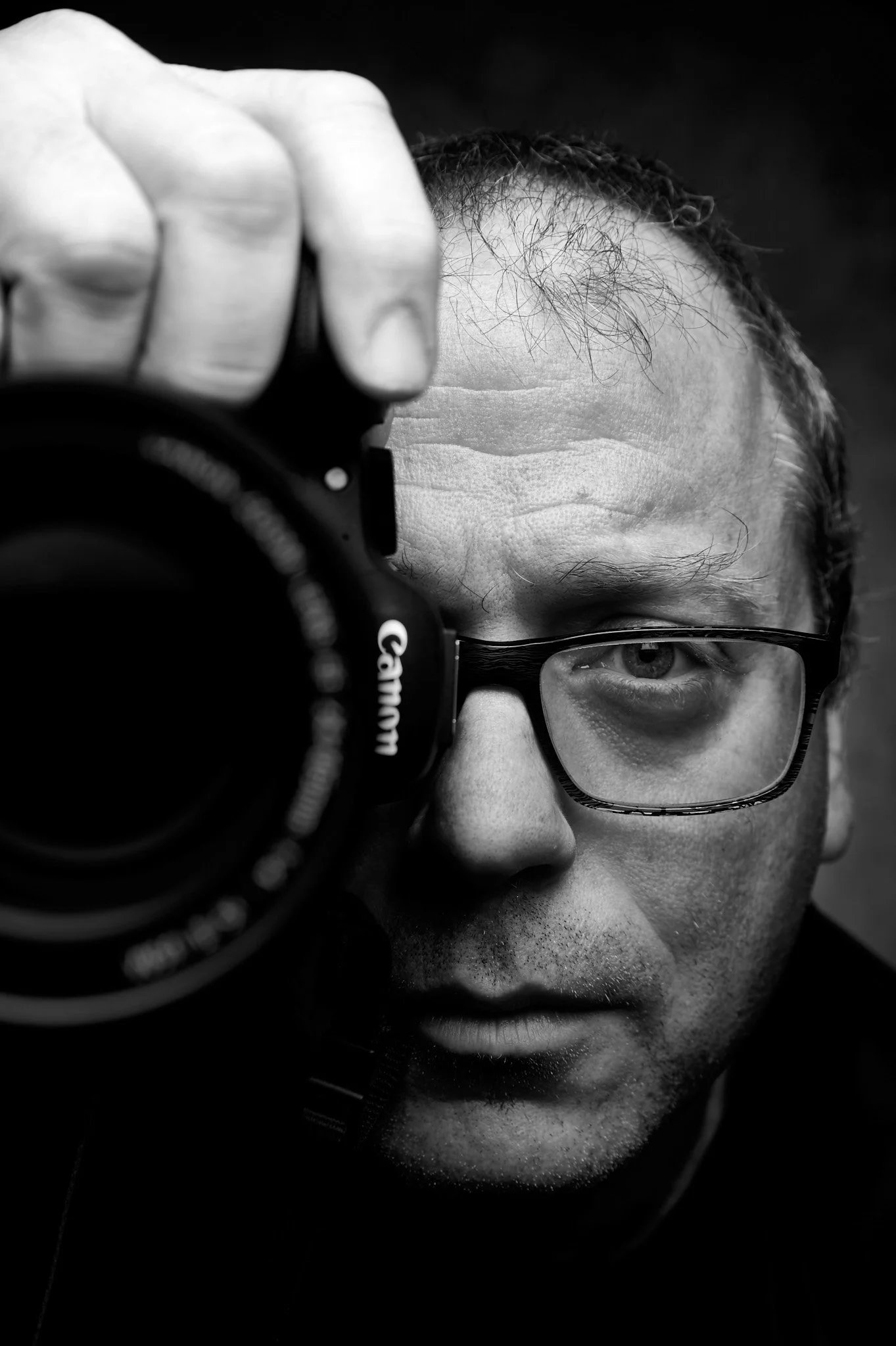 A man looks through a camera's viewfinder, with his face close to the camera and glasses on, in a black and white close-up shot.