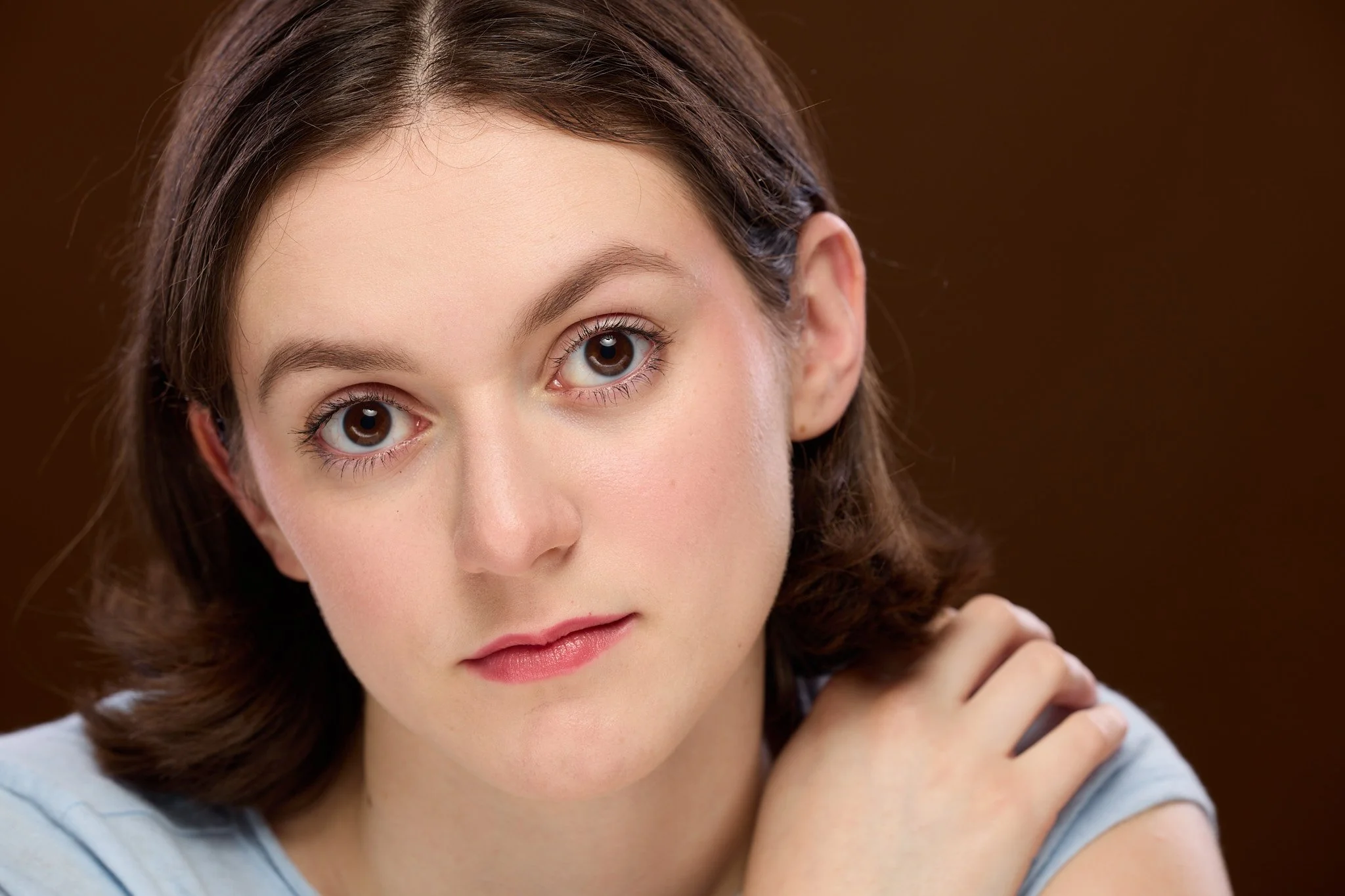 Close-up of a woman with brown hair, blue eyes, and fair skin, wearing a light blue top, with a neutral background.
