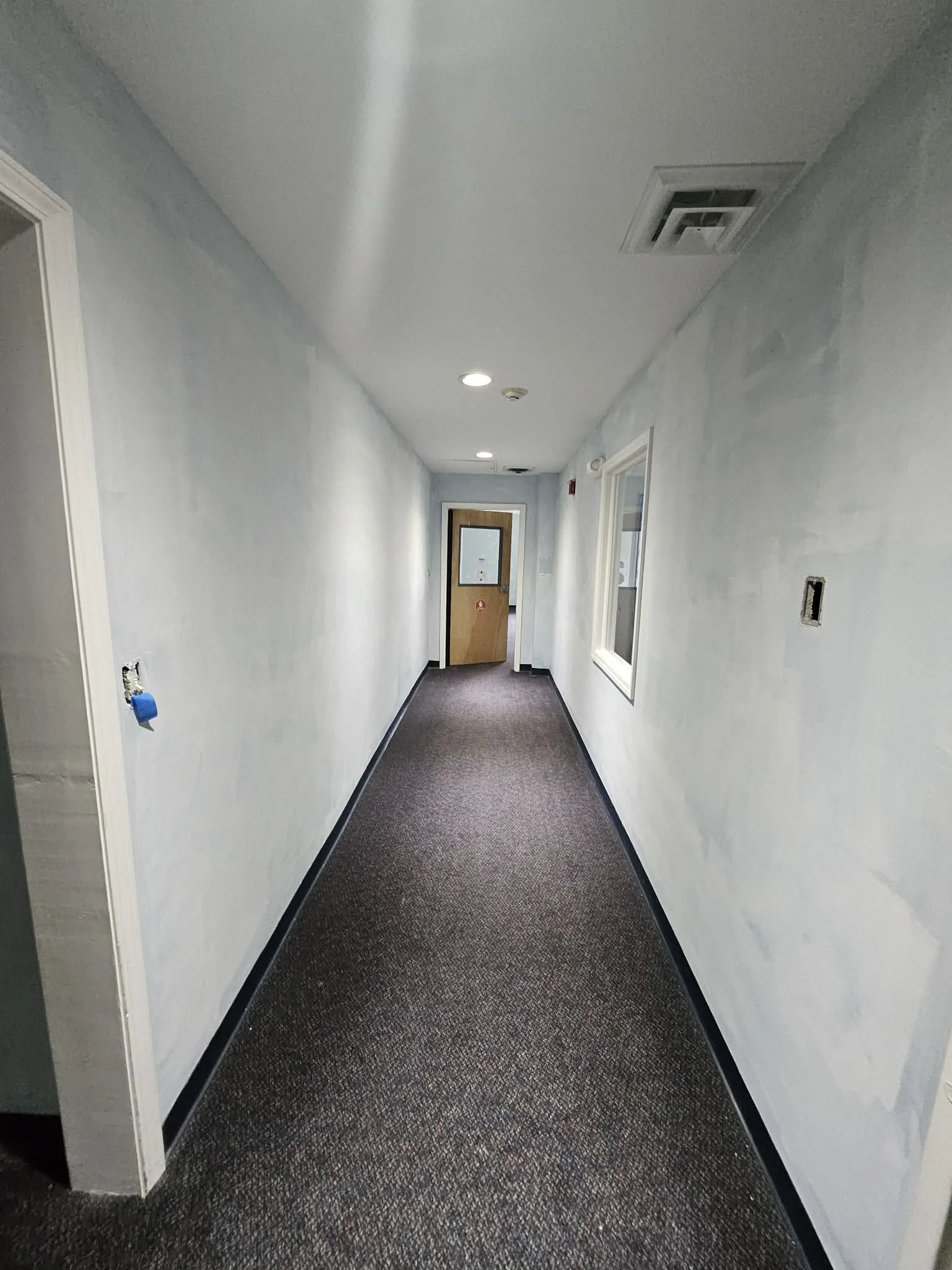 Long, narrow hallway with light blue walls, black window frames, a brown door at the end, and a brown carpeted floor.