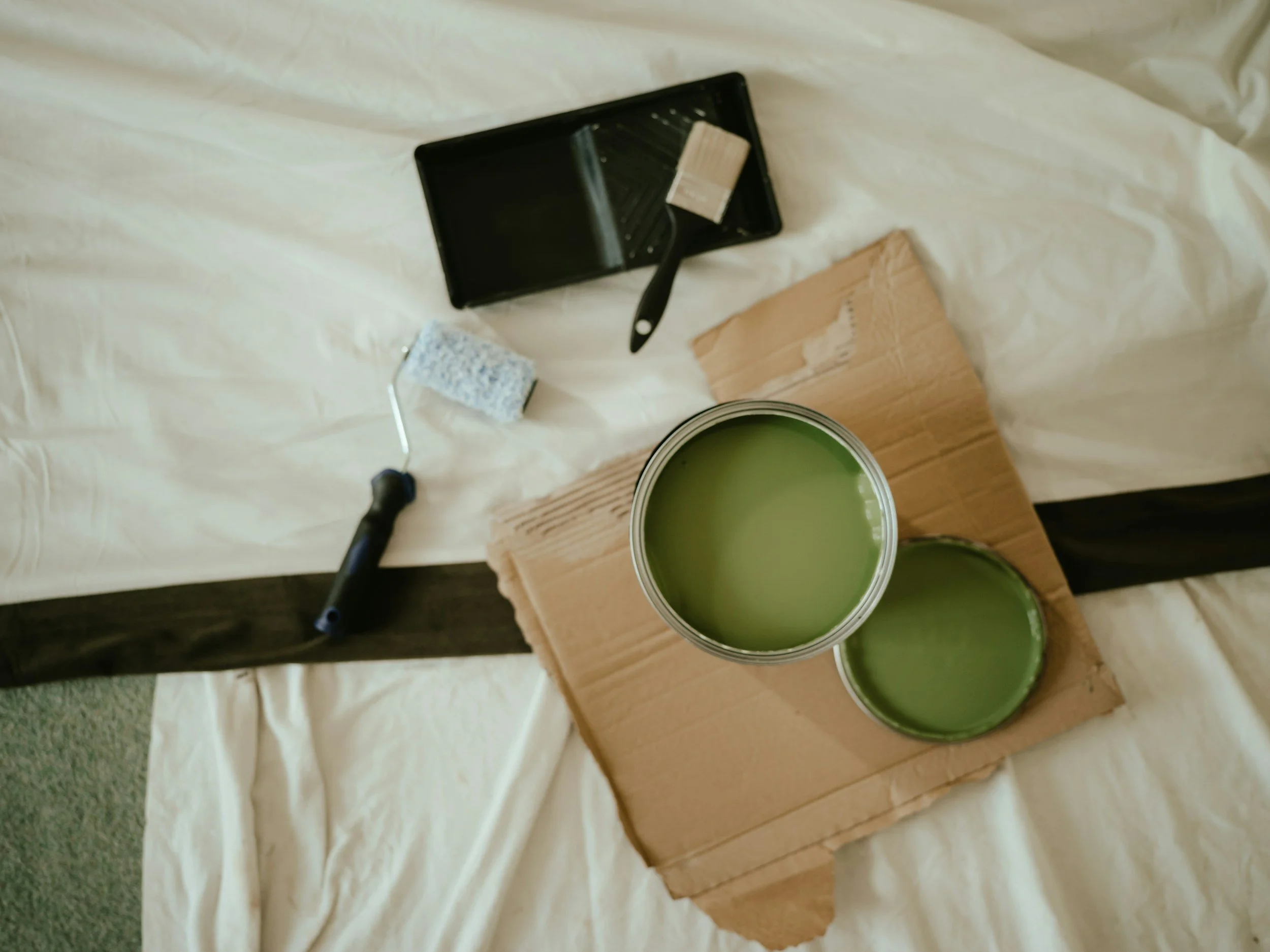 A flat lay of painting supplies on a white cloth, including a small paint roller, a paint tray with light beige paint, a cardboard piece, and two cans of green paint on a wooden board.