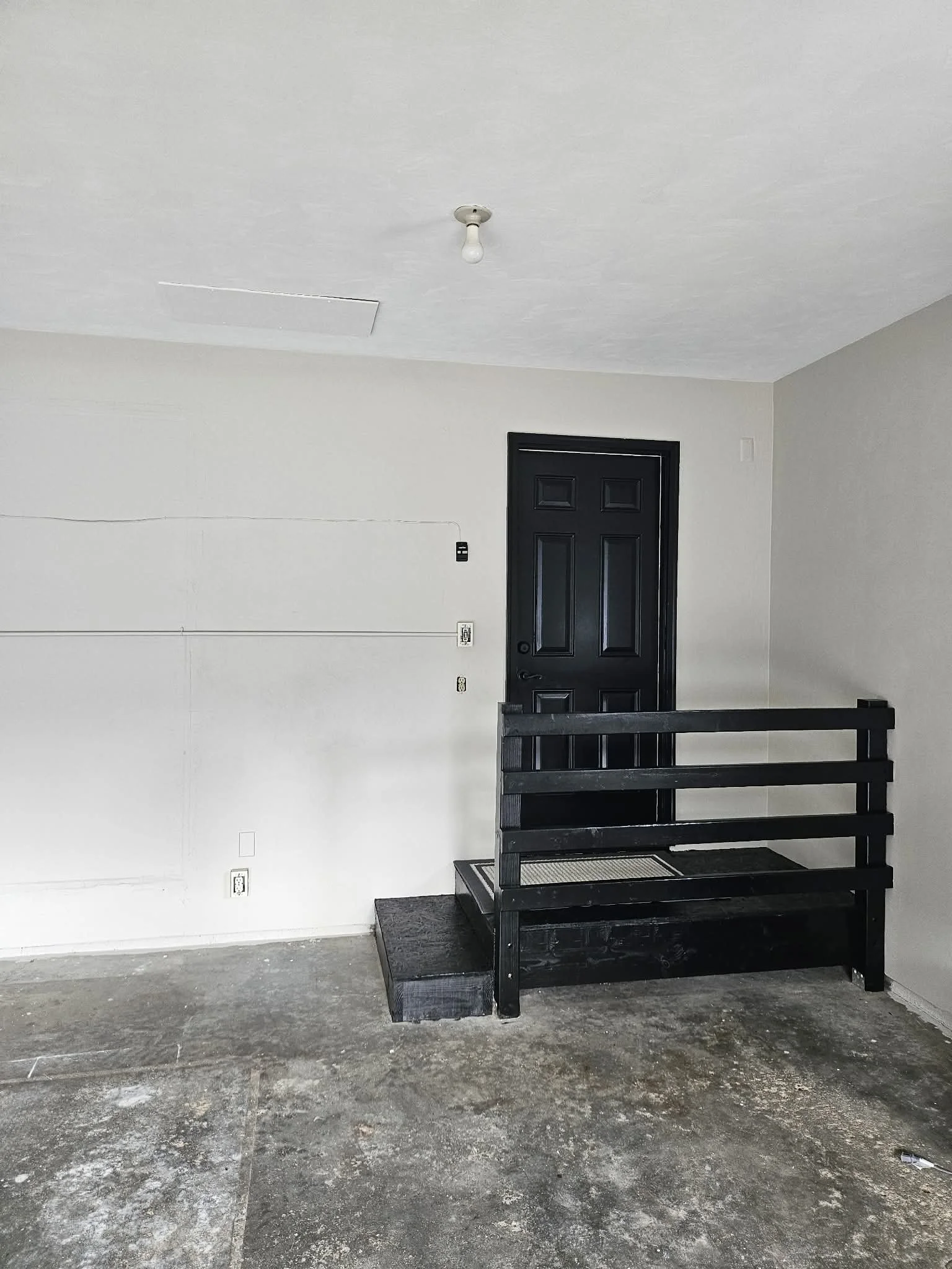 Interior of a room with a black door, black staircase, and a white wall with electrical outlets; the floor appears unfinished.
