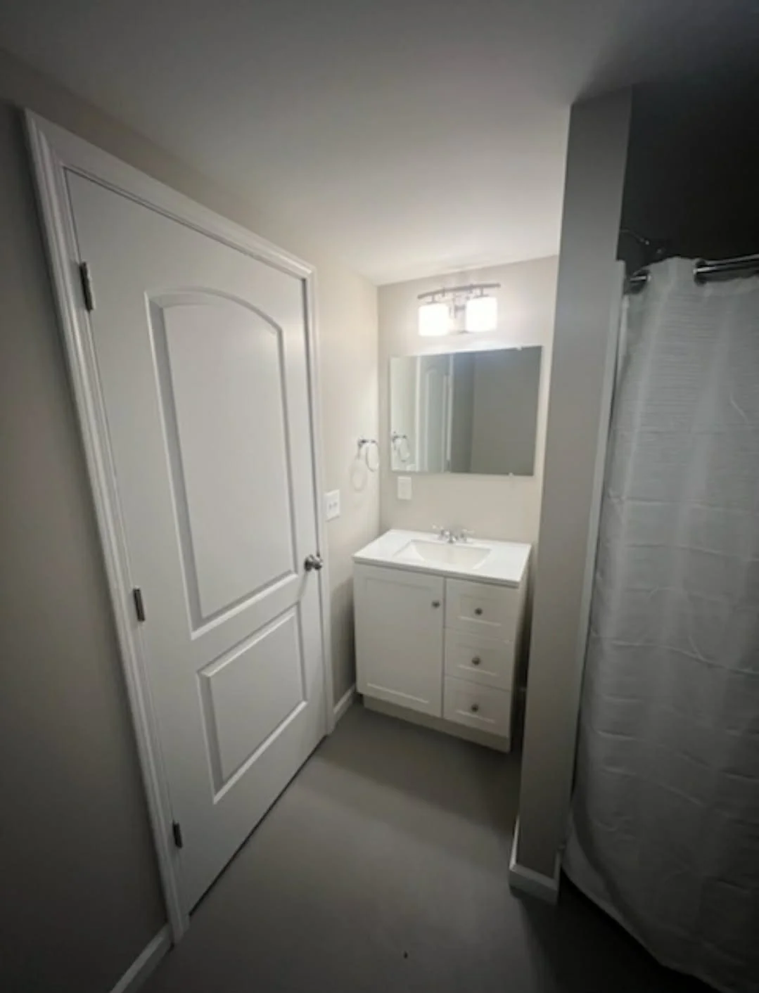 A small bathroom with a white vanity, mirror, and a window above. A closed door is on the left, and part of a shower curtain is on the right.