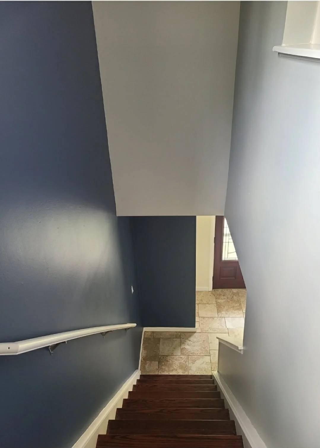 View looking down a staircase from above, showing dark wooden stairs, blue walls, a white handrail, and a tiled entrance area with a door at the bottom.