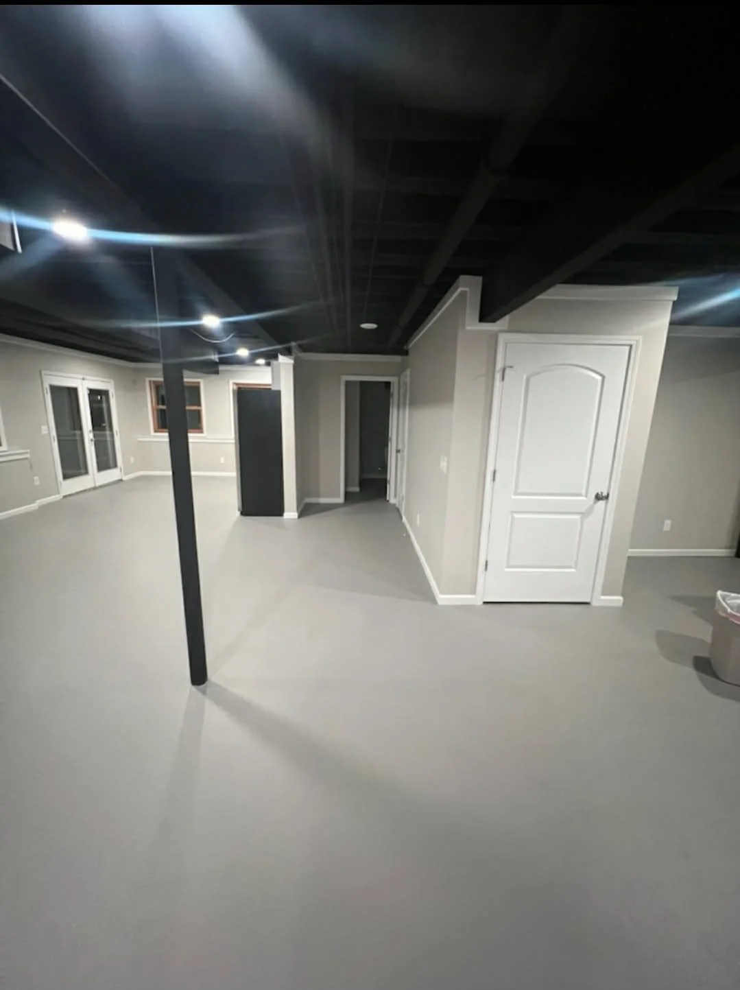 Empty basement room with light gray walls, light gray flooring, white doors, and black ceiling beams, illuminated by ceiling lights.