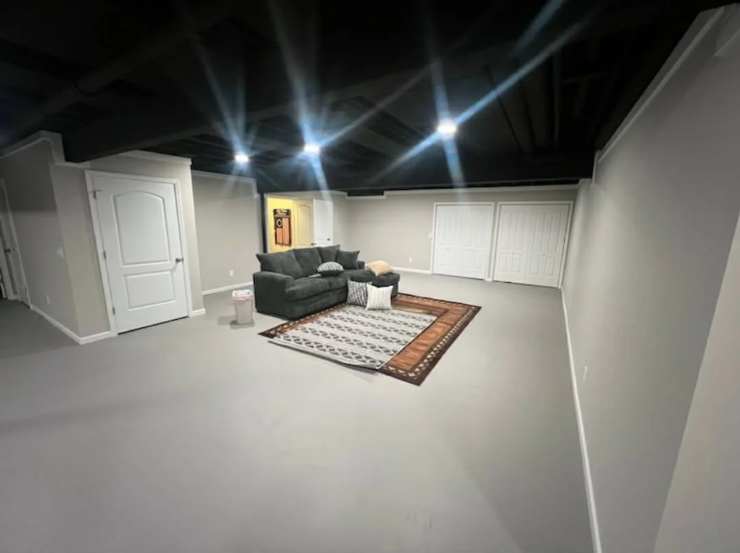 Empty basement with a dark ceiling, gray walls, white doors, a gray sofa with pillows, a patterned rug, and a small trash can. Overhead lights are on.