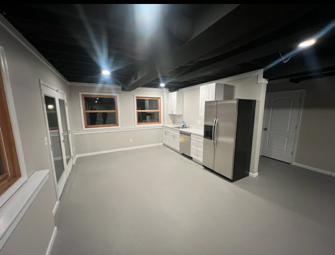 Empty modern kitchen with white cabinets, stainless steel refrigerator, dishwasher, black ceiling beams, three windows, and gray flooring.