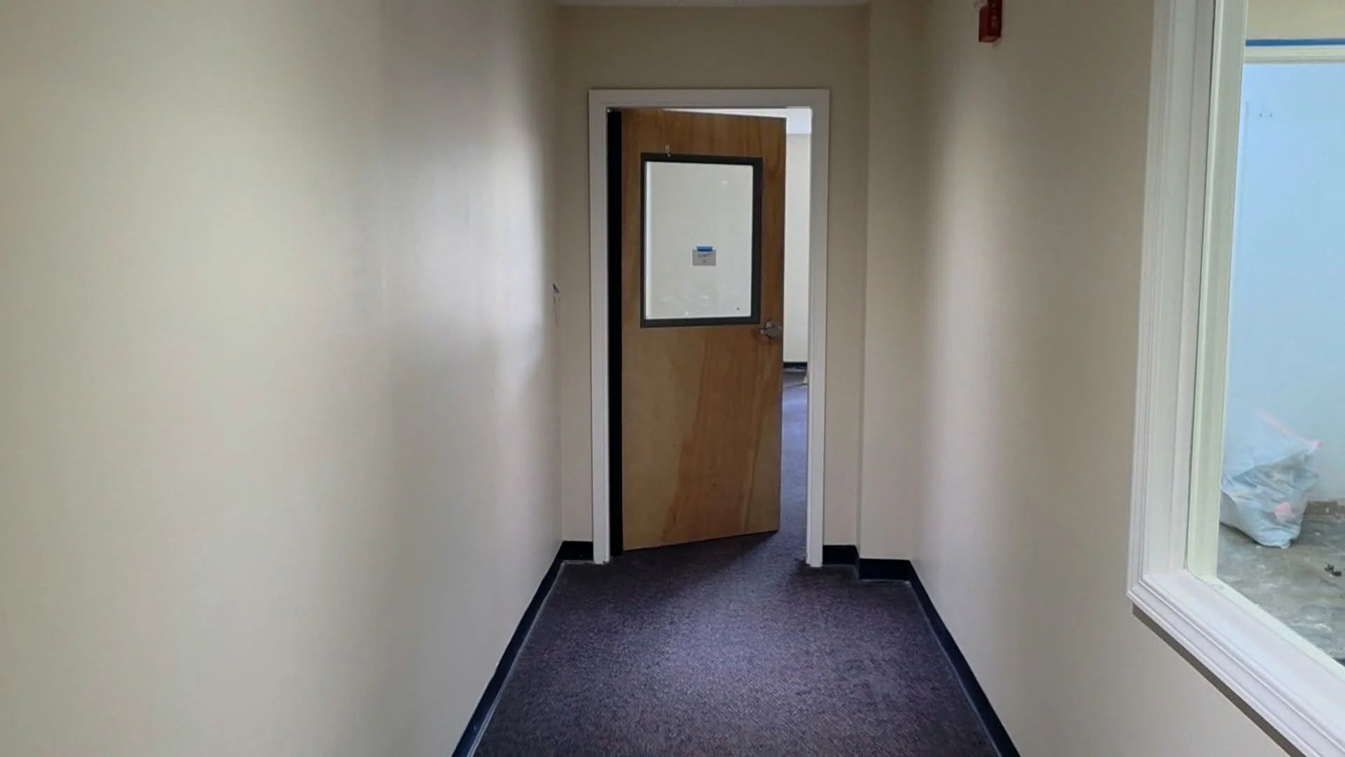 Empty hallway with beige walls, dark-colored carpet, a wooden door slightly open leading to a room with blue walls, and a window on the right side showing a plastic bag outside.