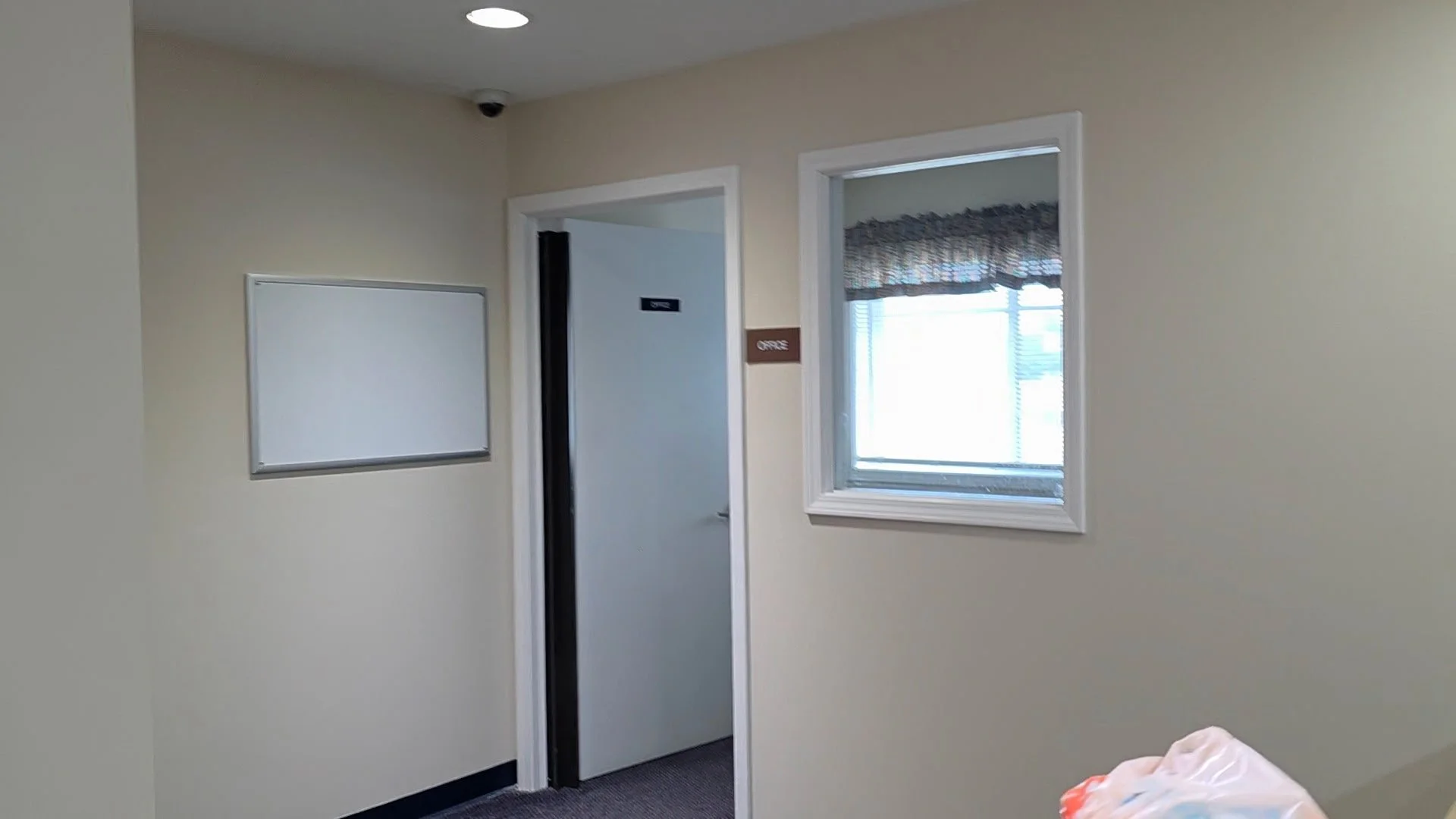 Interior view of a beige-colored room with a window with blinds, a whiteboard on the wall, and a doorway leading to another room or hallway.