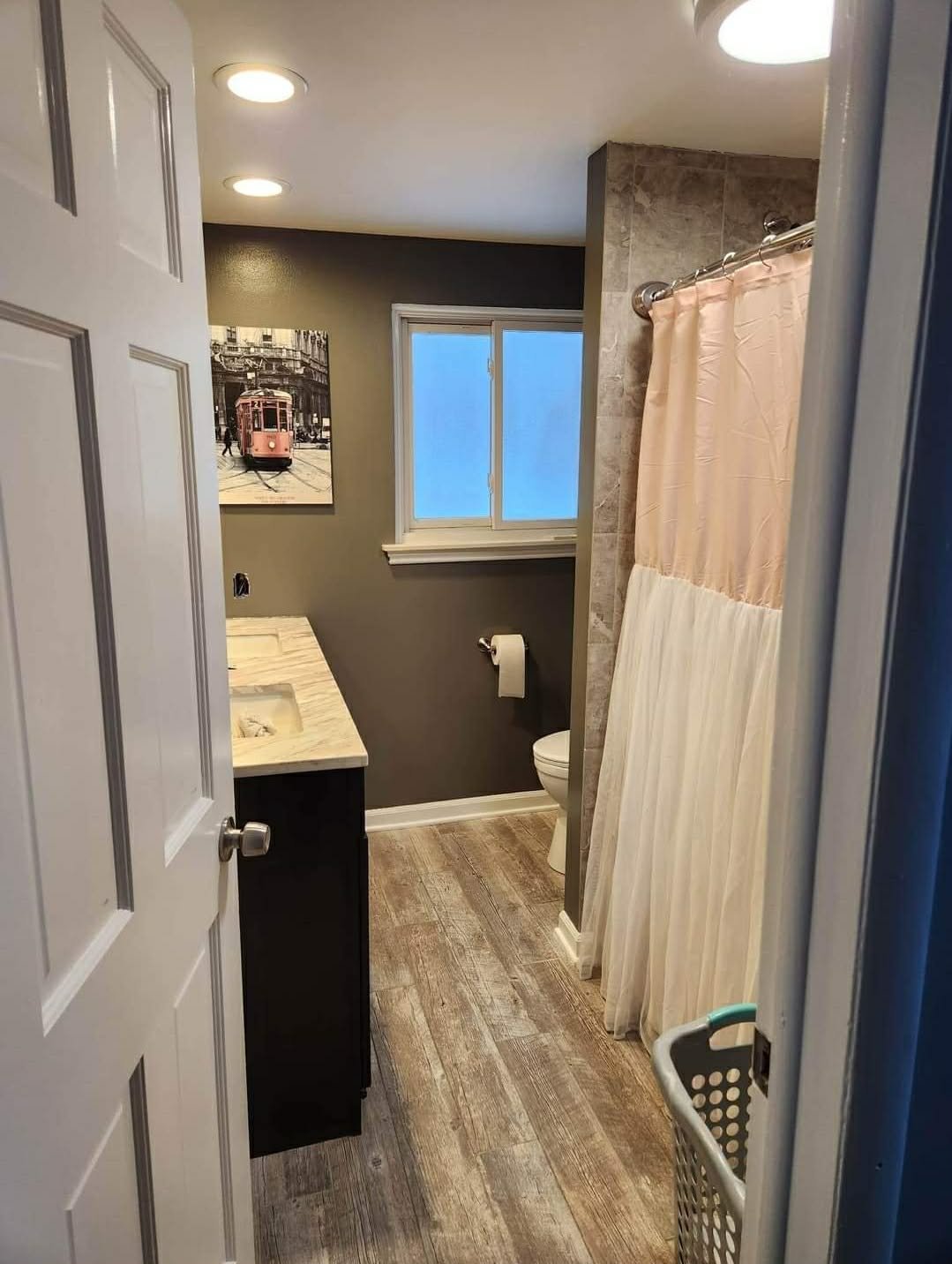 Small bathroom with gray walls, wooden flooring, a window, a toilet, a shower with a pink and white curtain, and a black vanity with a beige marble countertop.