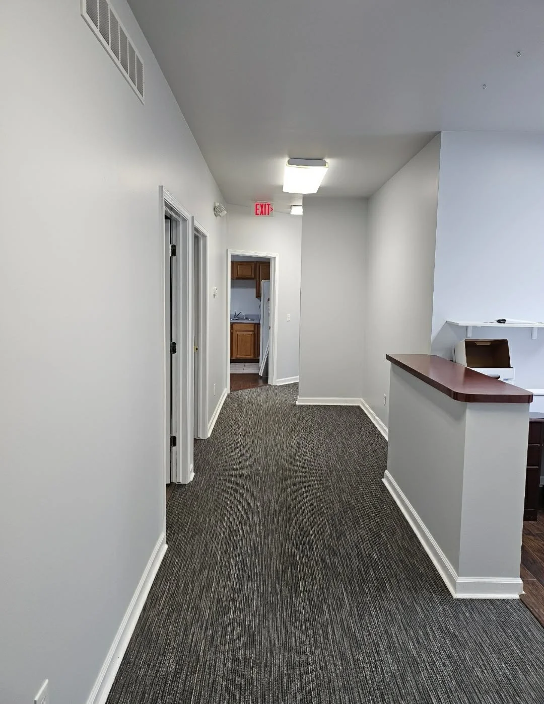 Empty hallway with gray walls, carpeted floor, ceiling light, and an open door leading to a room with wooden cabinets.