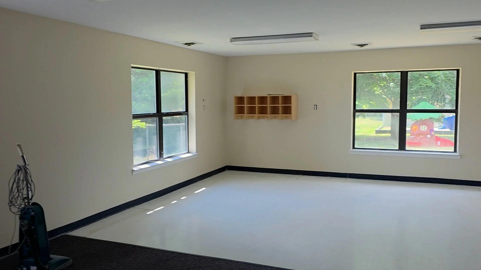 Empty room with light-colored walls, two large windows showing outdoor greenery and a playground, a small wooden shelf on one wall, and a vacuum cleaner in the corner.