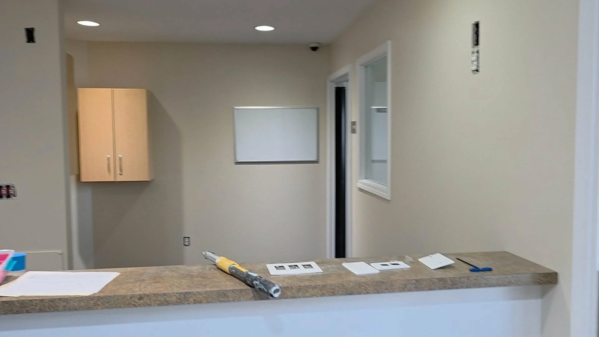 Interior view of a room under construction or renovation, with beige walls, a small wall-mounted cabinet on the left, a window on the right, and a countertop in the foreground with tools and papers.