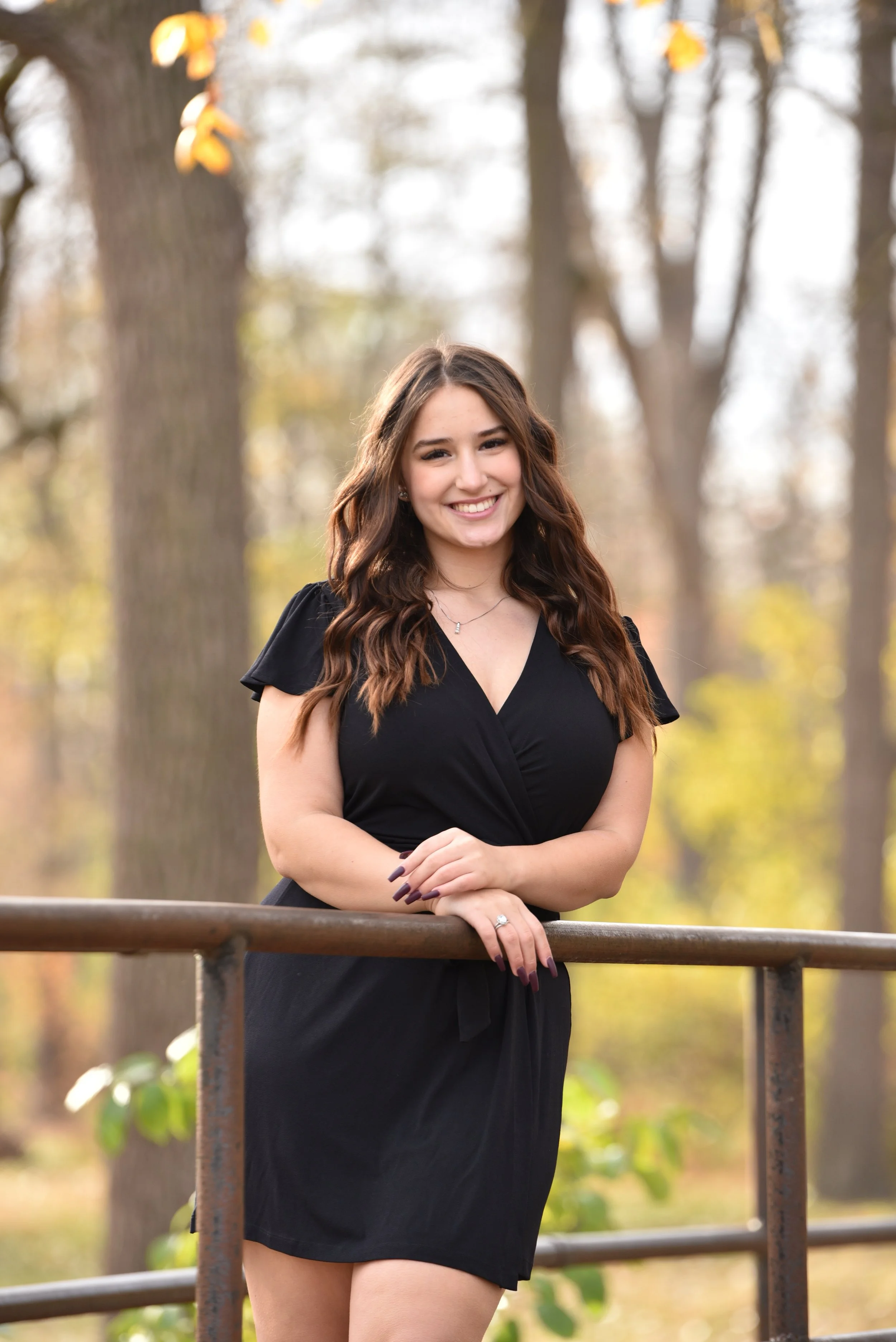 Young woman with long wavy brown hair smiling outdoors in a black dress, leaning on a metal railing with trees in the background.