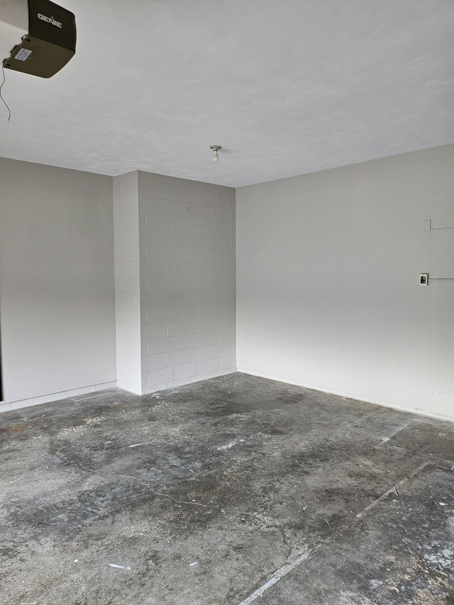 Empty room with unfinished concrete floor, gray painted walls, a small brick column, and a ceiling with a single exposed light bulb. There is an open electrical outlet and a wall-mounted box near the ceiling.