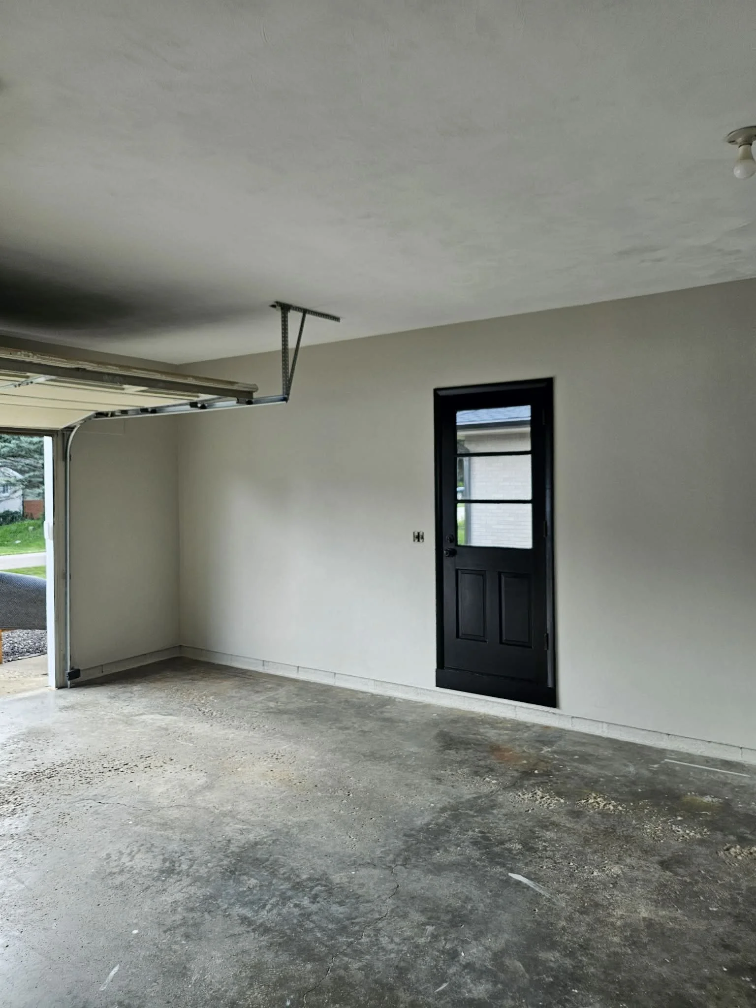 Interior of a garage with an unfinished concrete floor, a black door with a window, a partially open garage door, and an overhead garage door opener.