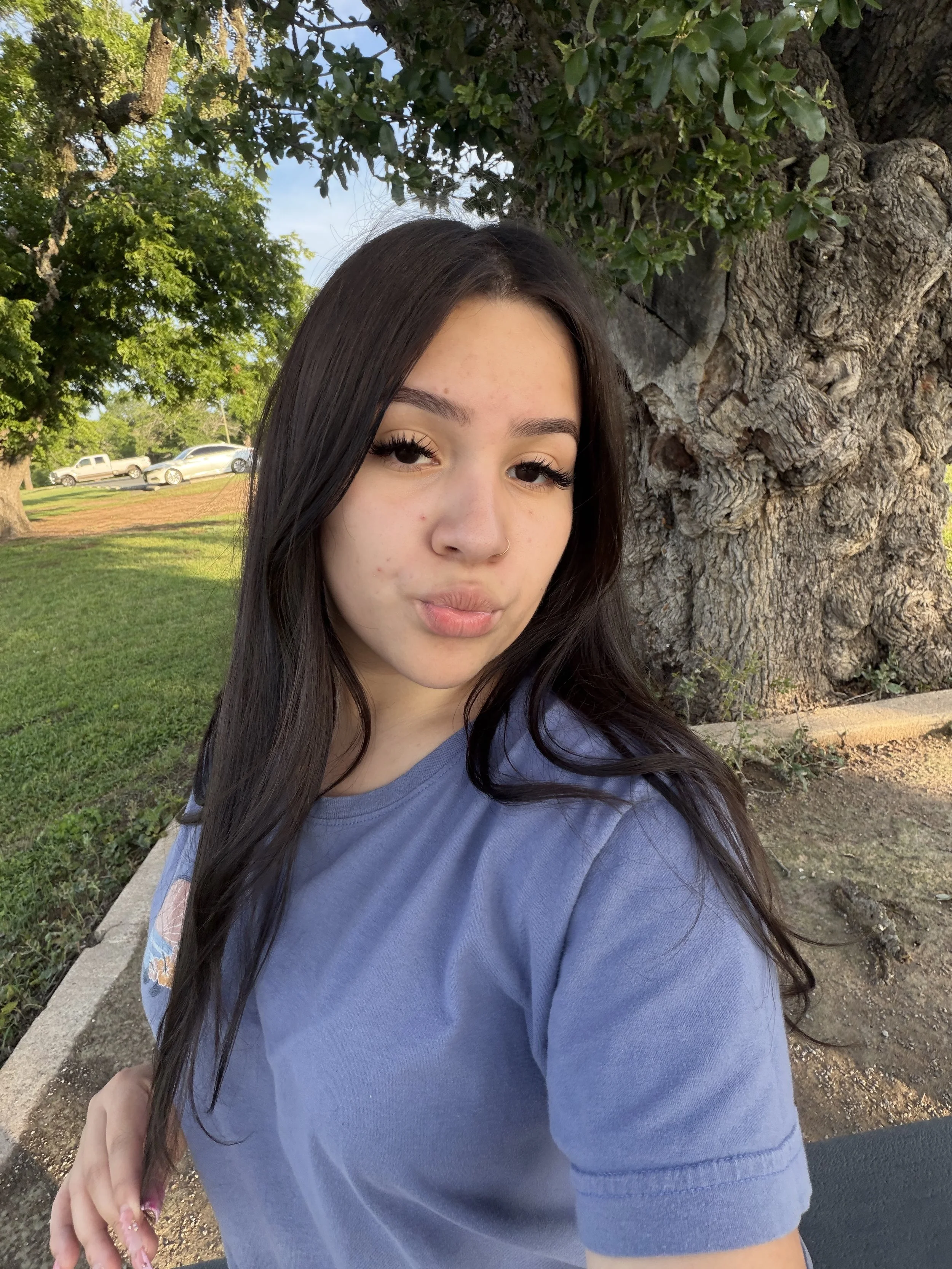 A young woman with long dark hair and a nose piercing taking a selfie outdoors near a large tree with green leaves, in a park or green space on a sunny day.