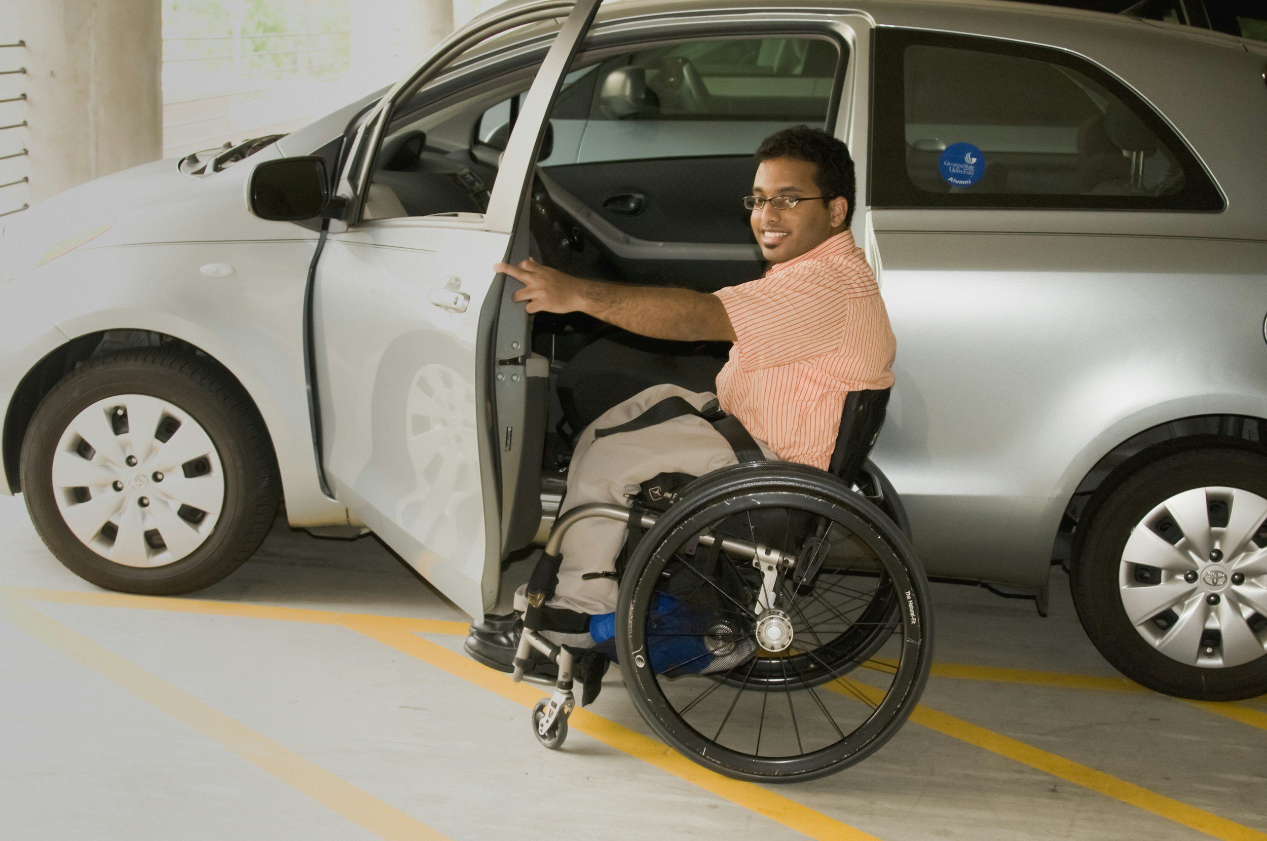 Young man in a wheelchair smiling, sitting next to a silver car in an indoor parking garage, with a sticker on the car window that says 'Alumni'.