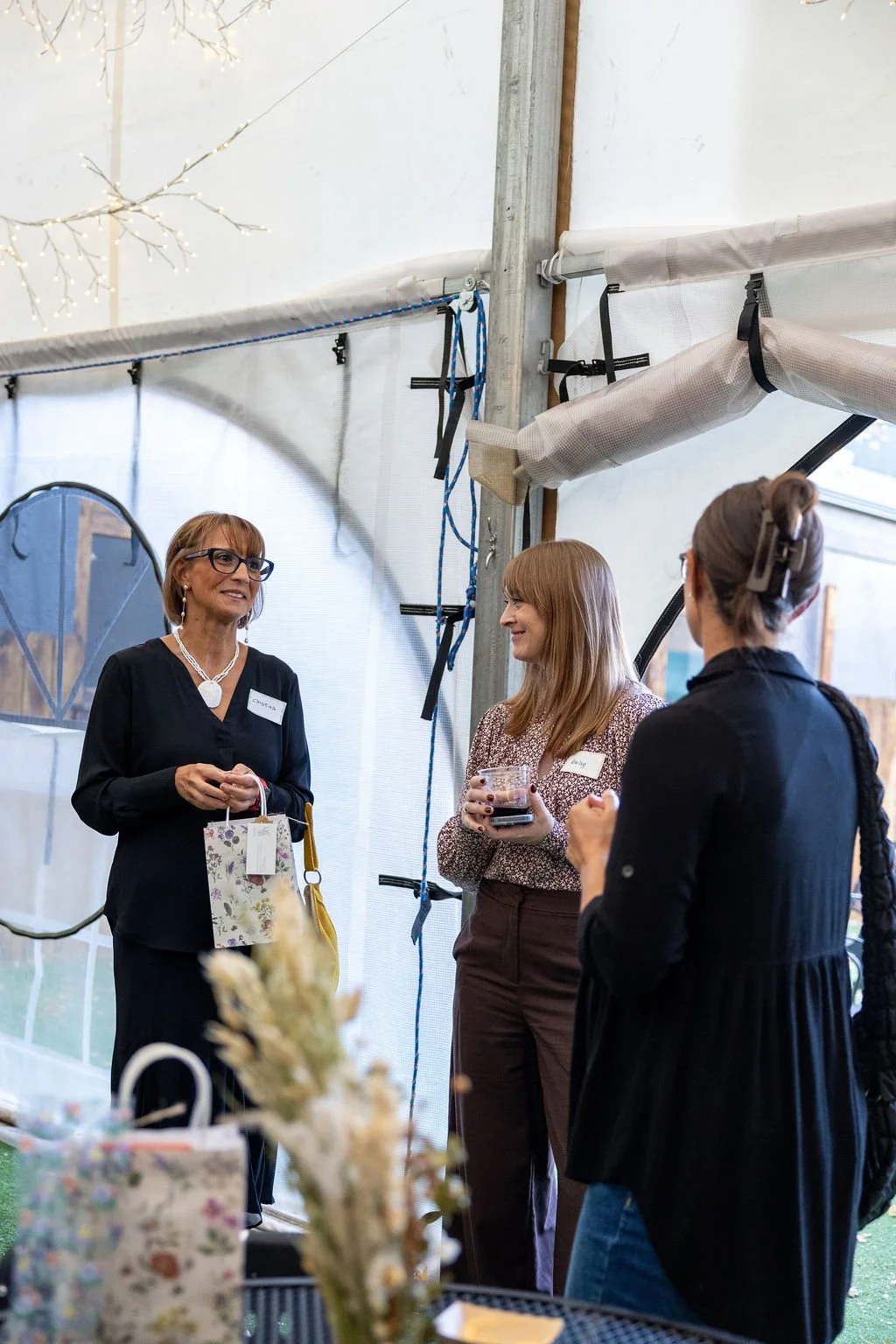 Three women conversing at an indoor event, with a rustic tent background, decorative lighting, and floral arrangements in the foreground.