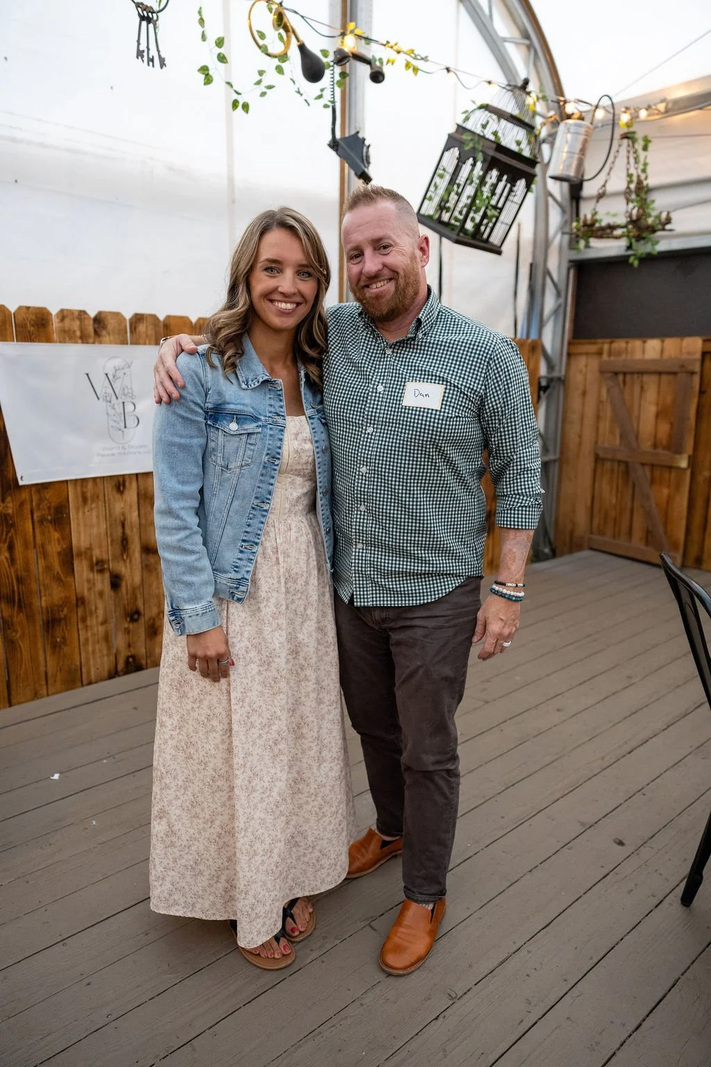 A smiling woman in a light-colored dress and denim jacket stands next to a smiling man with a beard and short hair, wearing a checkered shirt, dark pants, and brown shoes. They are in a wooden-fenced indoor setting with hanging plants, decorations, a