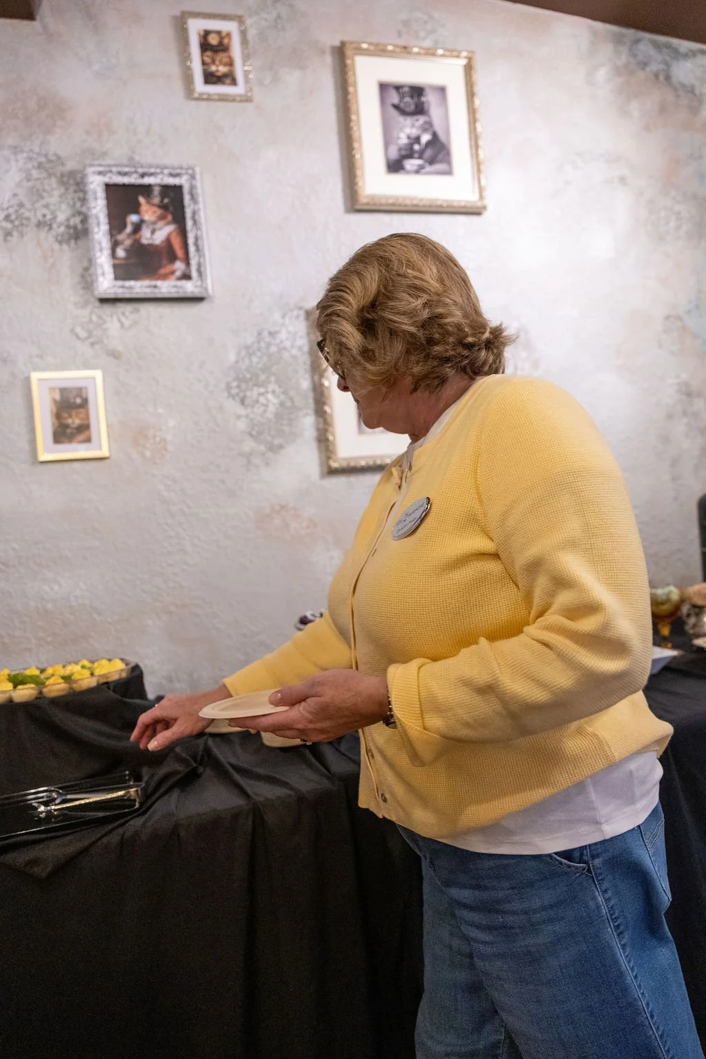 A woman in a yellow sweater is serving herself at a buffet table with small plates and food items, in a room decorated with framed pictures on the wall.