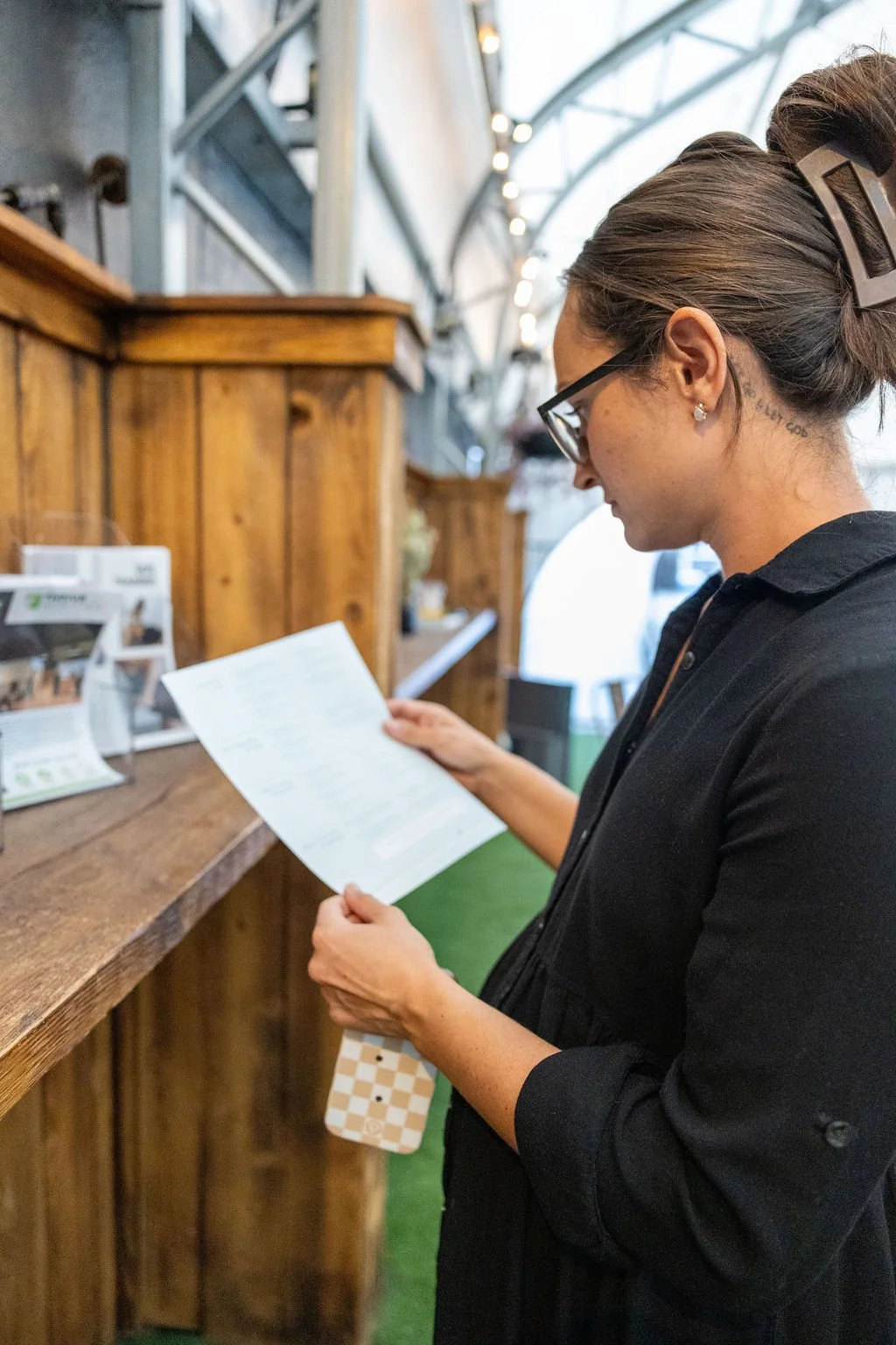 A woman with glasses and a black shirt looking at a menu at a wooden counter inside a building with a curved metal roof.