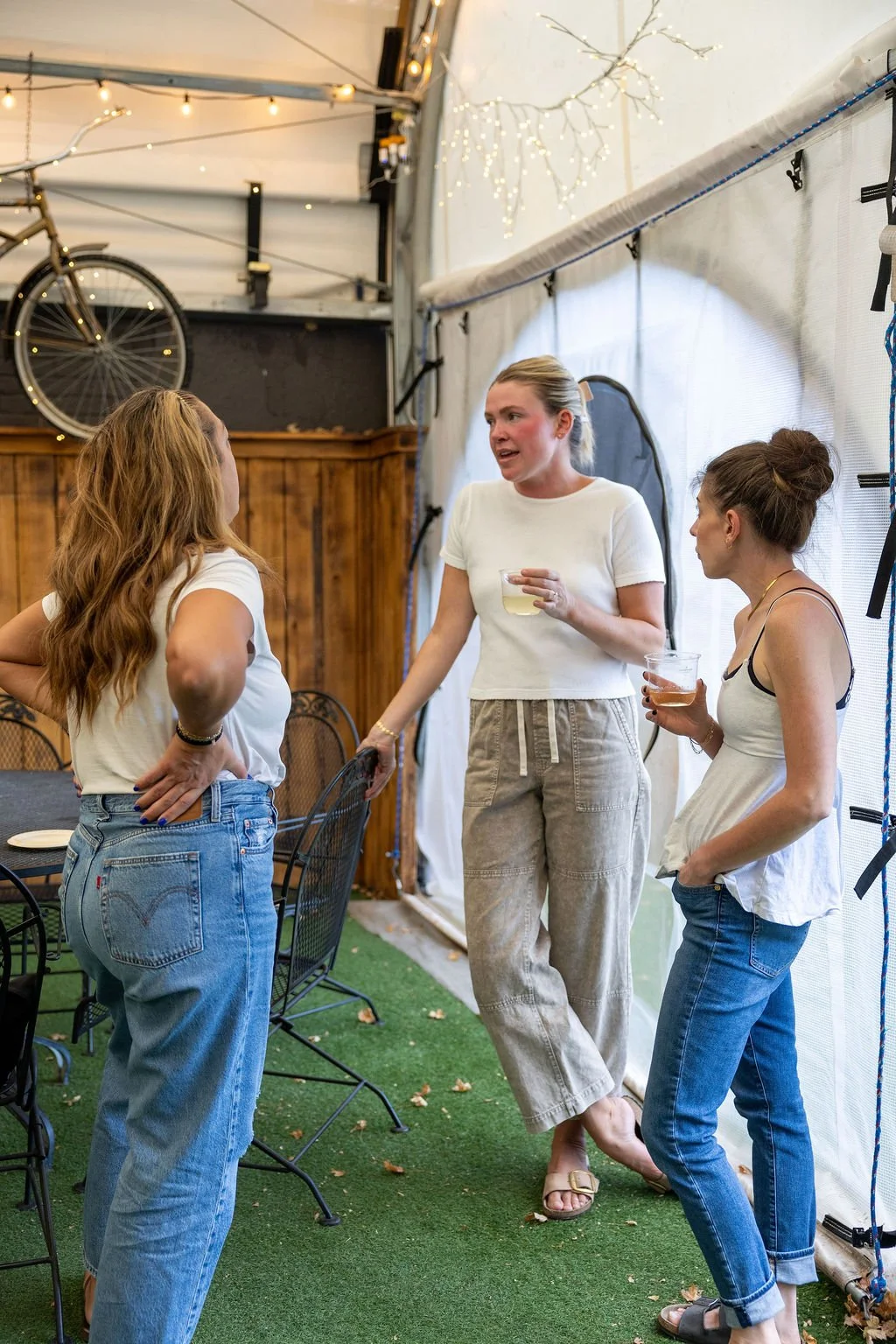 Three women having a conversation inside a room with string lights and a bicycle hanging on the wall, with a wooden panel behind them and some tables and chairs around.