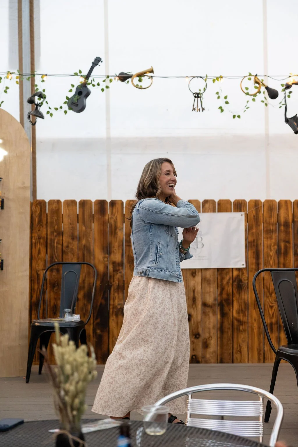 Woman in a denim jacket and floral skirt smiling and talking in a casual indoor space decorated with string lights, musical instruments, and wooden panels.