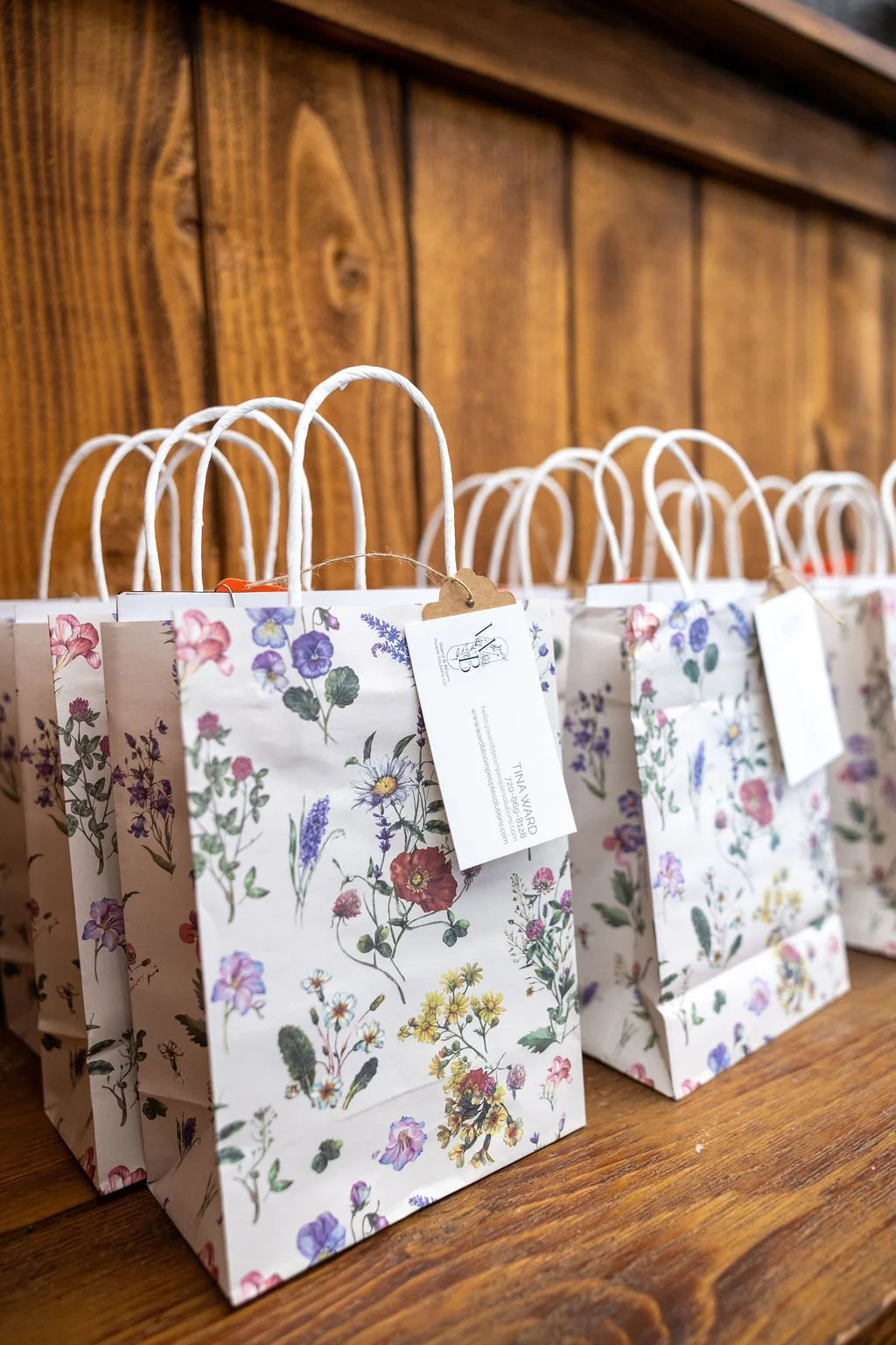 Multiple paper gift bags with floral patterns and white handles, arranged on a wooden surface against a wooden wall background.