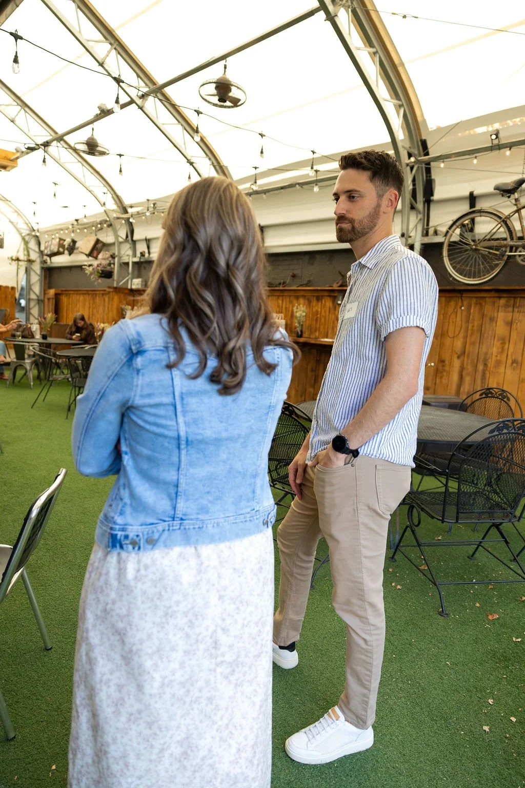 Two people having a conversation in a covered outdoor space with string lights and a wooden fence, with other people sitting at tables in the background.