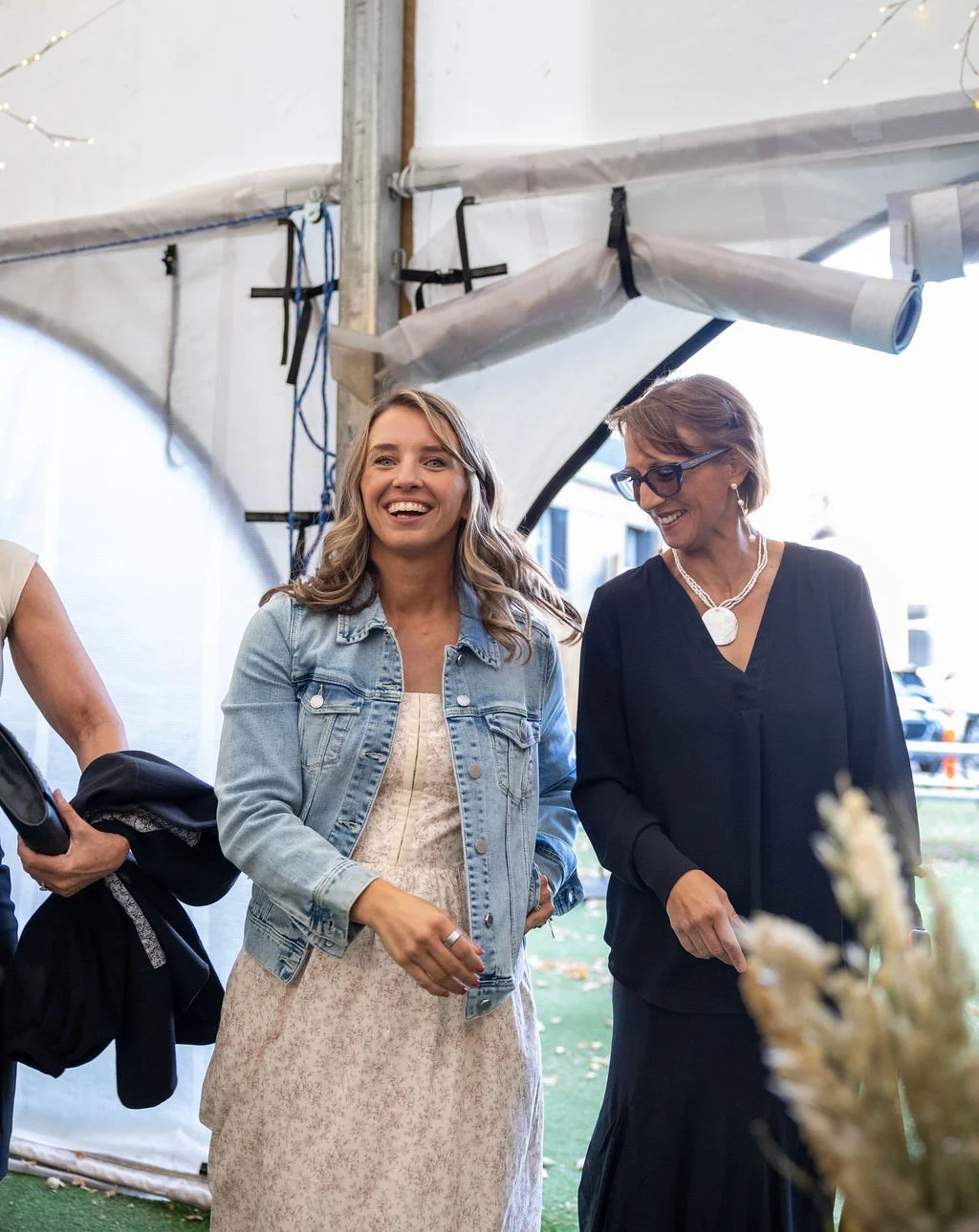 Two women smiling and walking together in an outdoor setting with a tent-like structure above them. One woman is wearing a denim jacket and dress, while the other is dressed in black with glasses and jewelry.