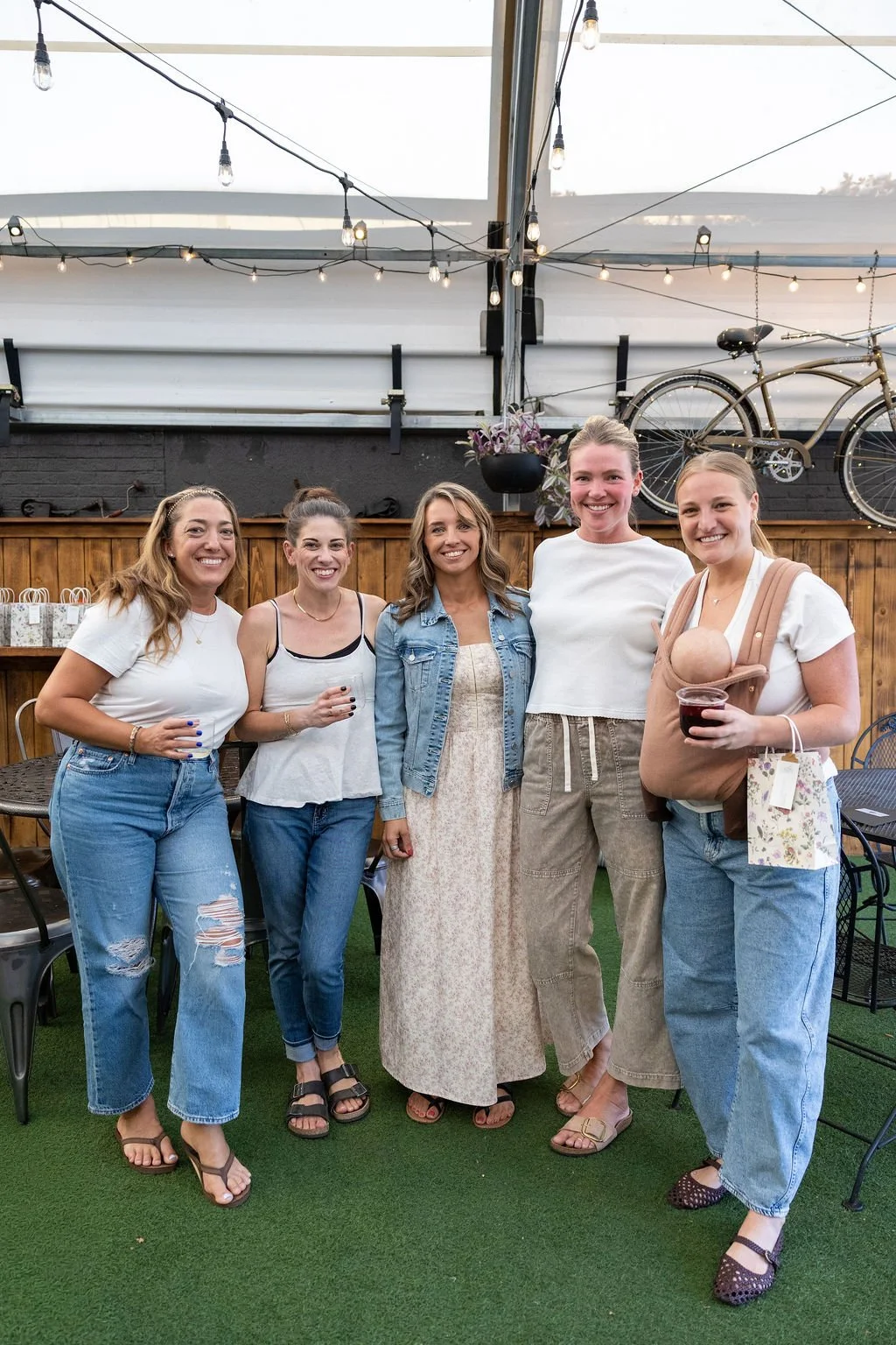 Group of five women standing in a row at an indoor gathering, with one holding a baby in a baby carrier and another holding a cup of wine, in a decorated venue with string lights and bicycles on the wall.