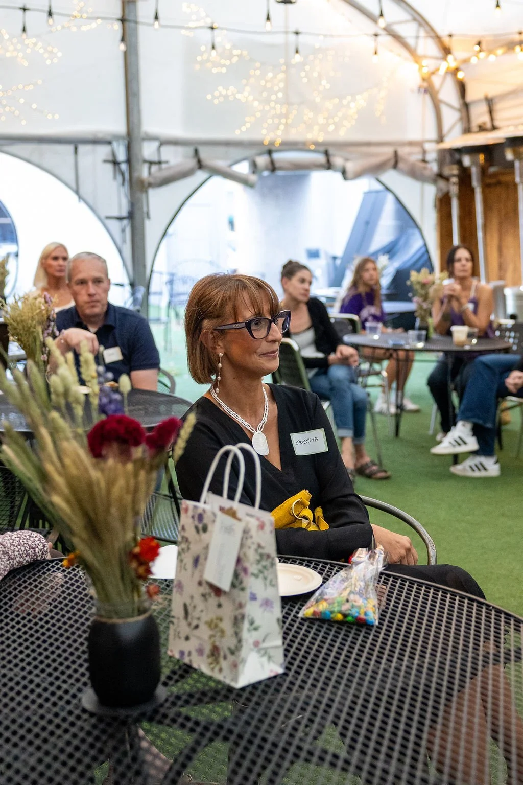 A woman with glasses and a name tag labeled 'Cristina' sitting at a table in a tented indoor event space, with other attendees in the background. The table has a vase with flowers, a gift bag, and a bag of colorful candies. Decorative string lights h