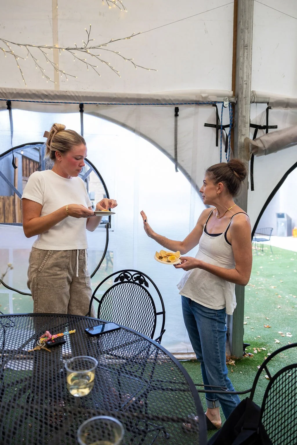 Two women are outdoors under a tent, one is holding a plate of food and the other is holding a small plate with a sandwich; they are engaged in a conversation, with the woman holding a sandwich showing a gesture. There are black metal chairs and a ta