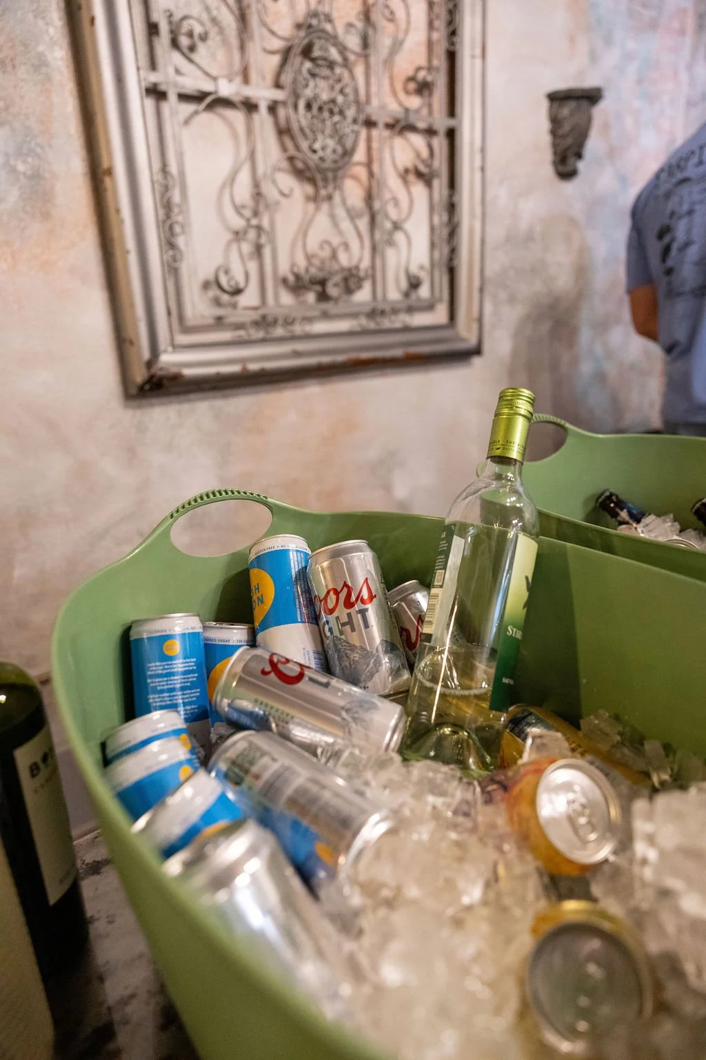 A green container filled with ice and empty beer cans, a beer bottle, and beverage cans, placed on a table in front of a textured wall and decorative metal gate.