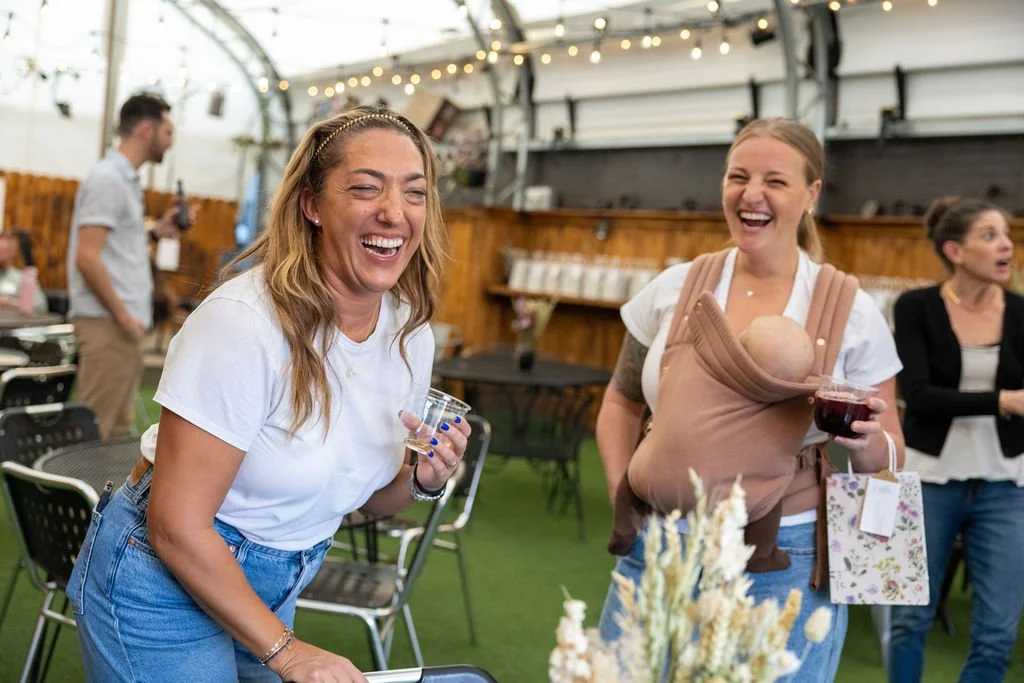 Two women are laughing and enjoying drinks at an indoor event, one wearing a baby carrier with a baby inside, and the other holding a glass of red wine.