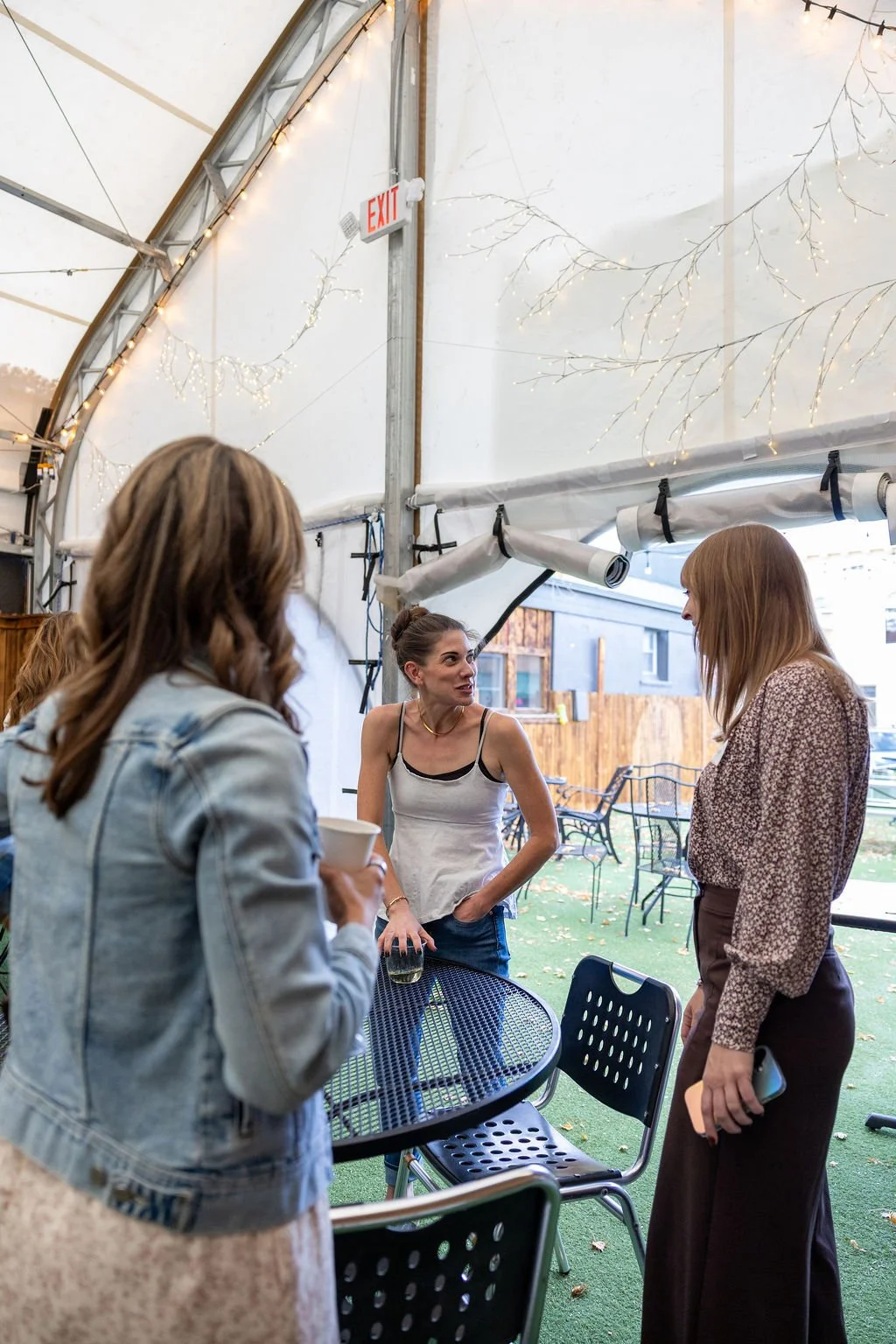 Group of women having a conversation at an outdoor event under a canopy with string lights and an exit sign.