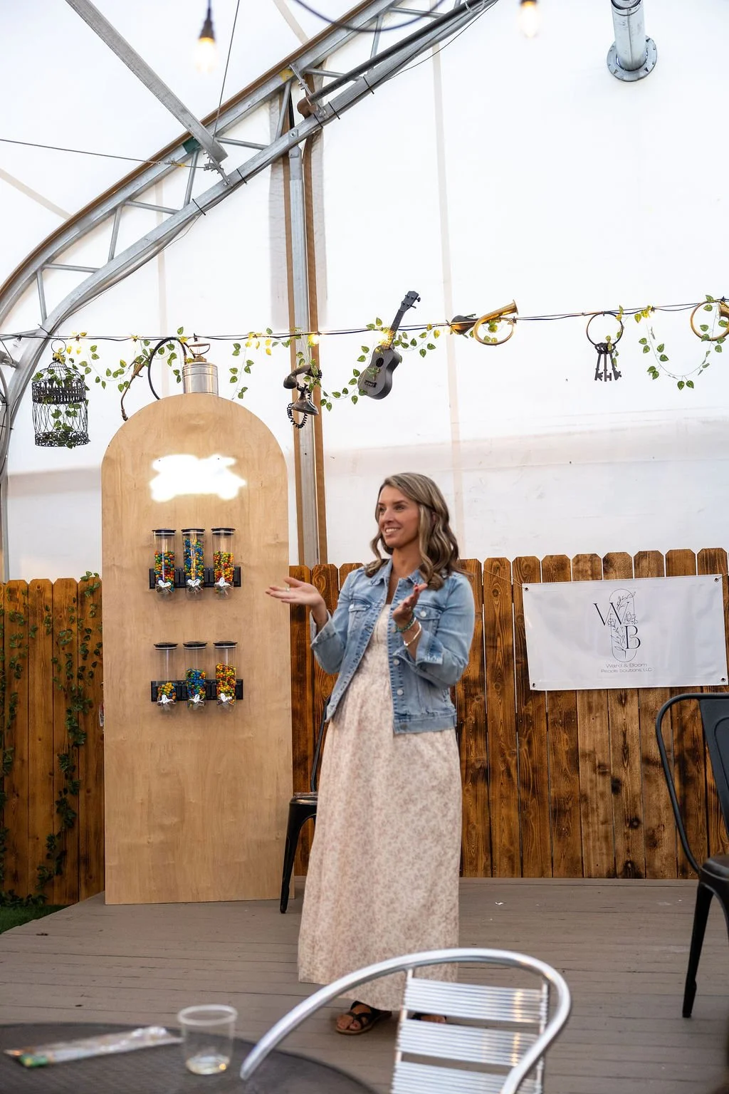 A smiling woman in a long dress and denim jacket talking at an indoor event with wooden fencing, hanging string lights, decorative keys, a small guitar, a birdcage, and a banner in the background.