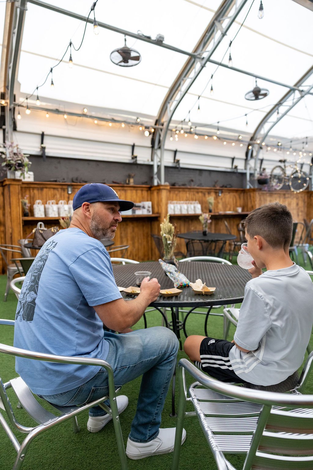 A man and boy sitting at a round outdoor table inside a greenhouse-style space, eating snacks and drinking water.