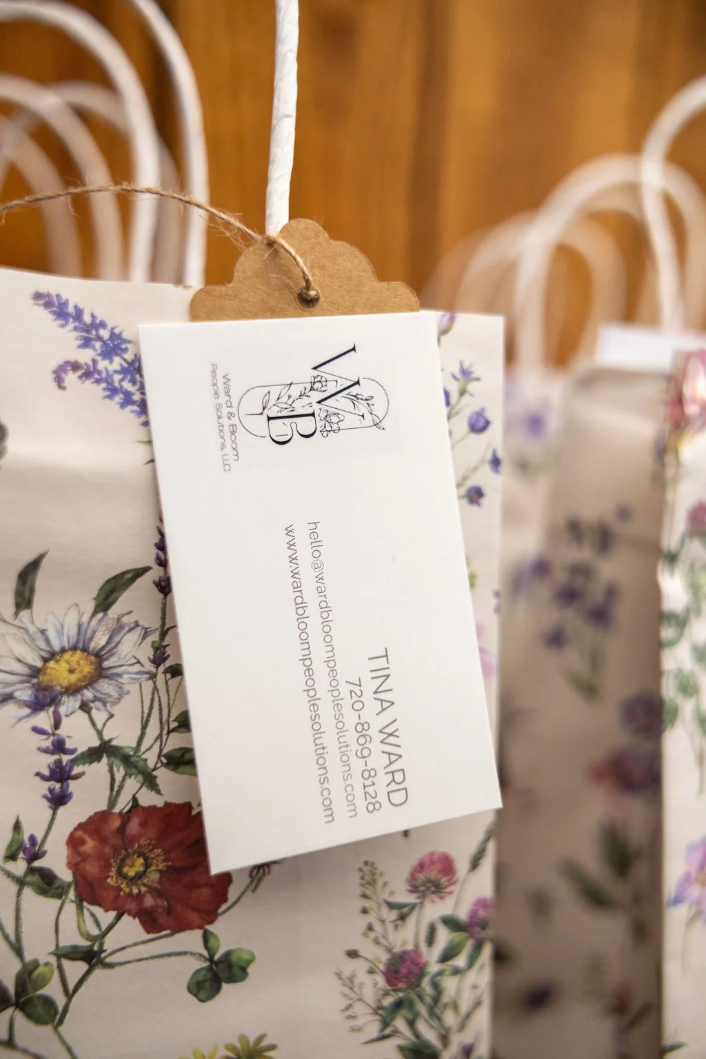 Close-up of a floral-patterned paper bag with a white tag attached, displaying contact information for Tina Ward and a floral illustration, with blurred additional similar bags in the background.
