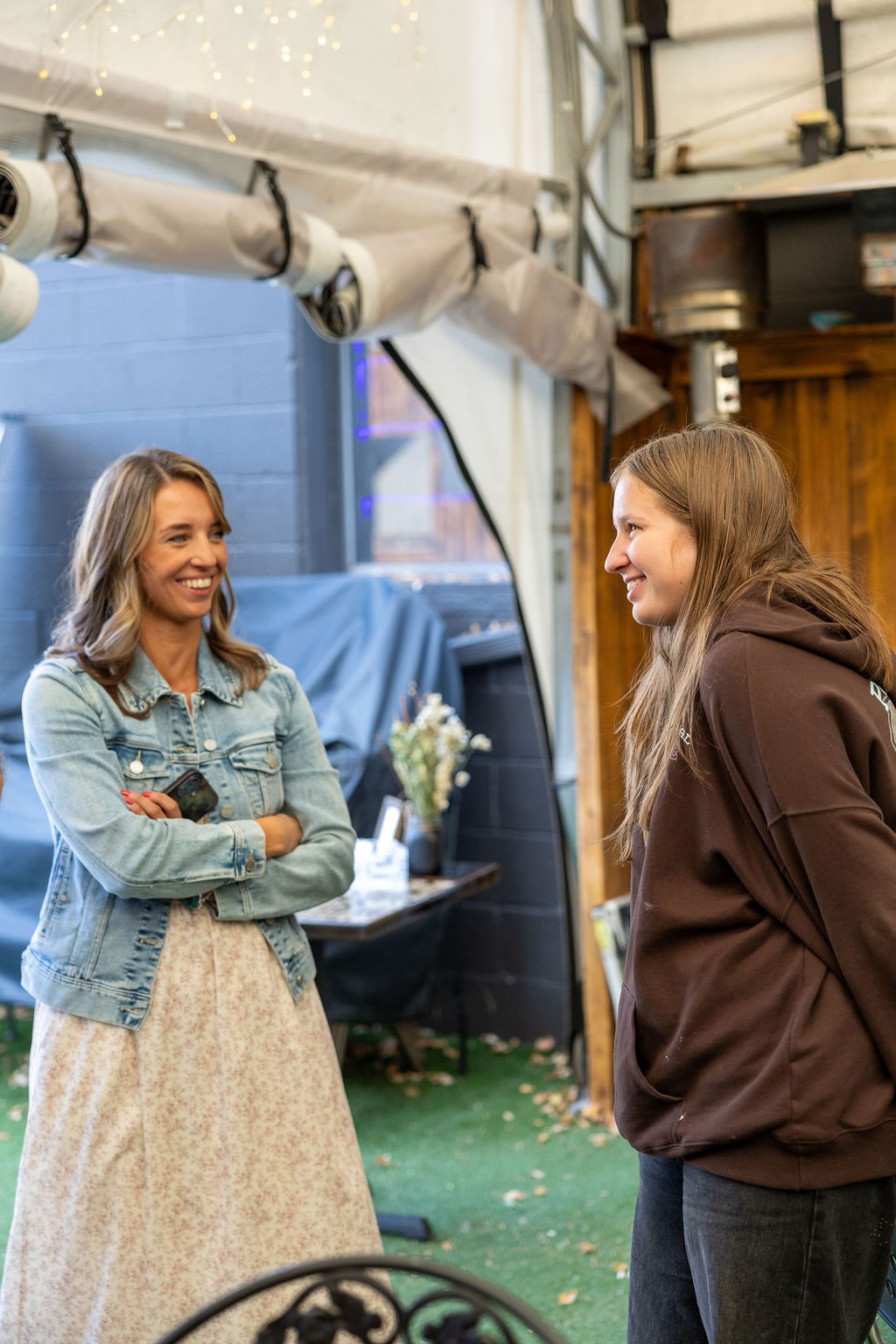 Two women are smiling and talking in a casual setting. One woman is wearing a denim jacket and a beige dress, and the other is wearing a brown hoodie. They are indoors with a background that includes a window, some plants, and wooden decor.