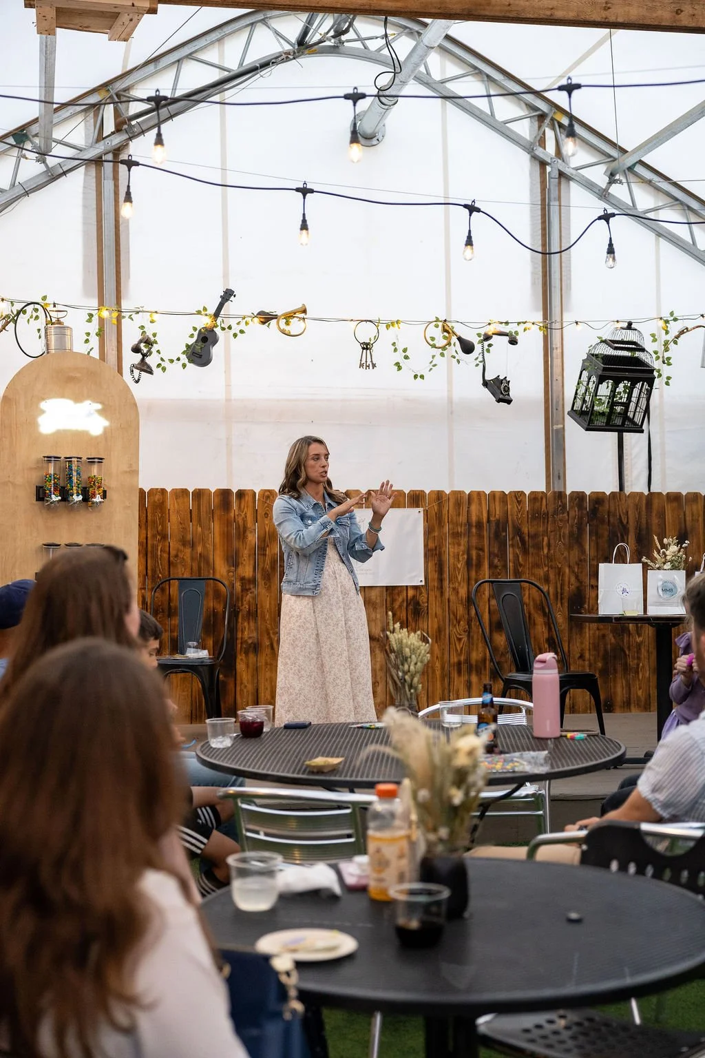 A woman standing and speaking in front of an audience in a greenhouse-style setting, decorated with hanging string lights, plants, and musical instruments.