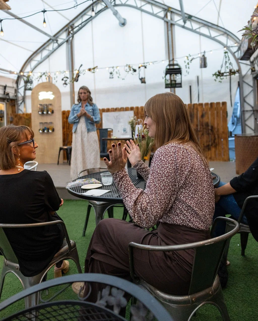 A woman with light brown hair and a patterned shirt sitting at an outdoor event, clapping, with another woman sitting nearby. A woman in a denim jacket and white dress stands on a stage in a greenhouse-like structure decorated with string lights and 