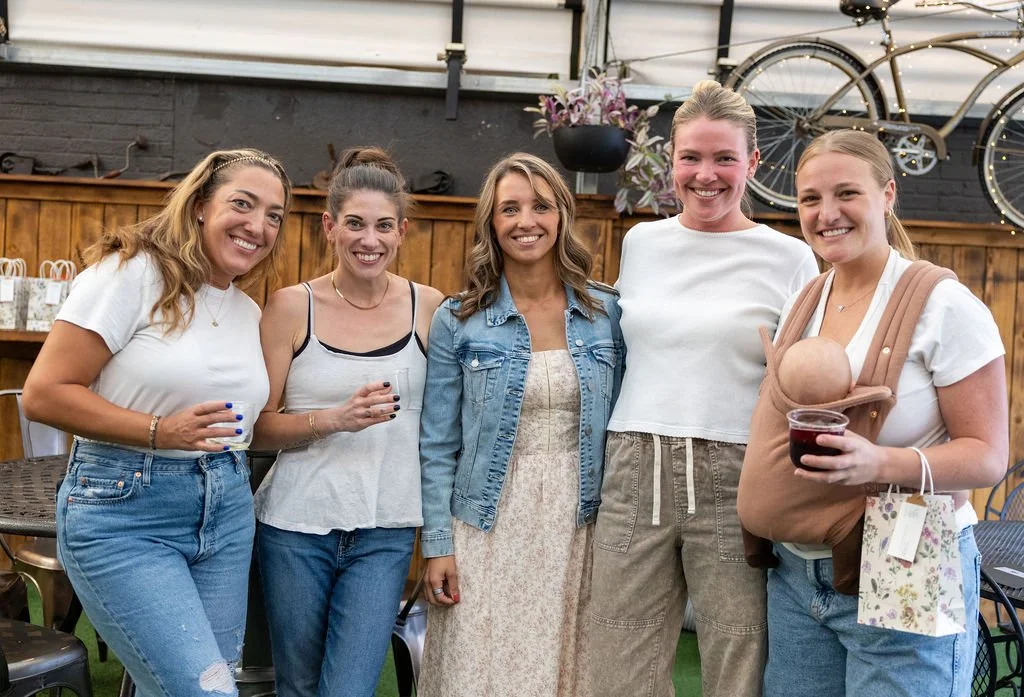 Group of five women standing together, smiling, in a cozy indoor setting with wooden walls and decorative bicycles in the background, one woman holding a baby in a carrier.